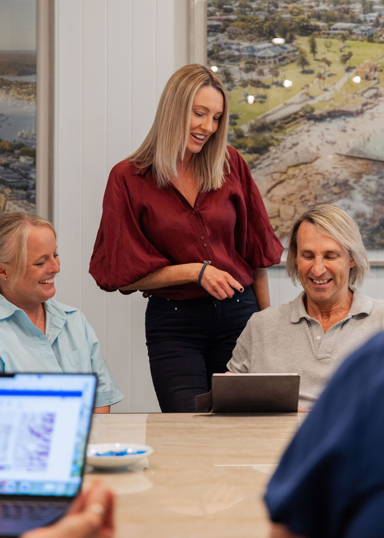 Three women smiling and collaborating around a table with laptops in a bright modern office meeting room.