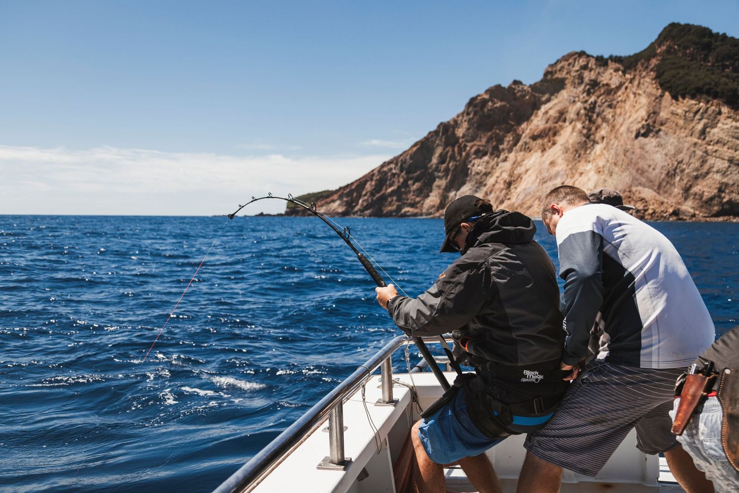 Two anglers fishing from a boat near rocky cliffs on deep blue ocean waters under a clear sky.