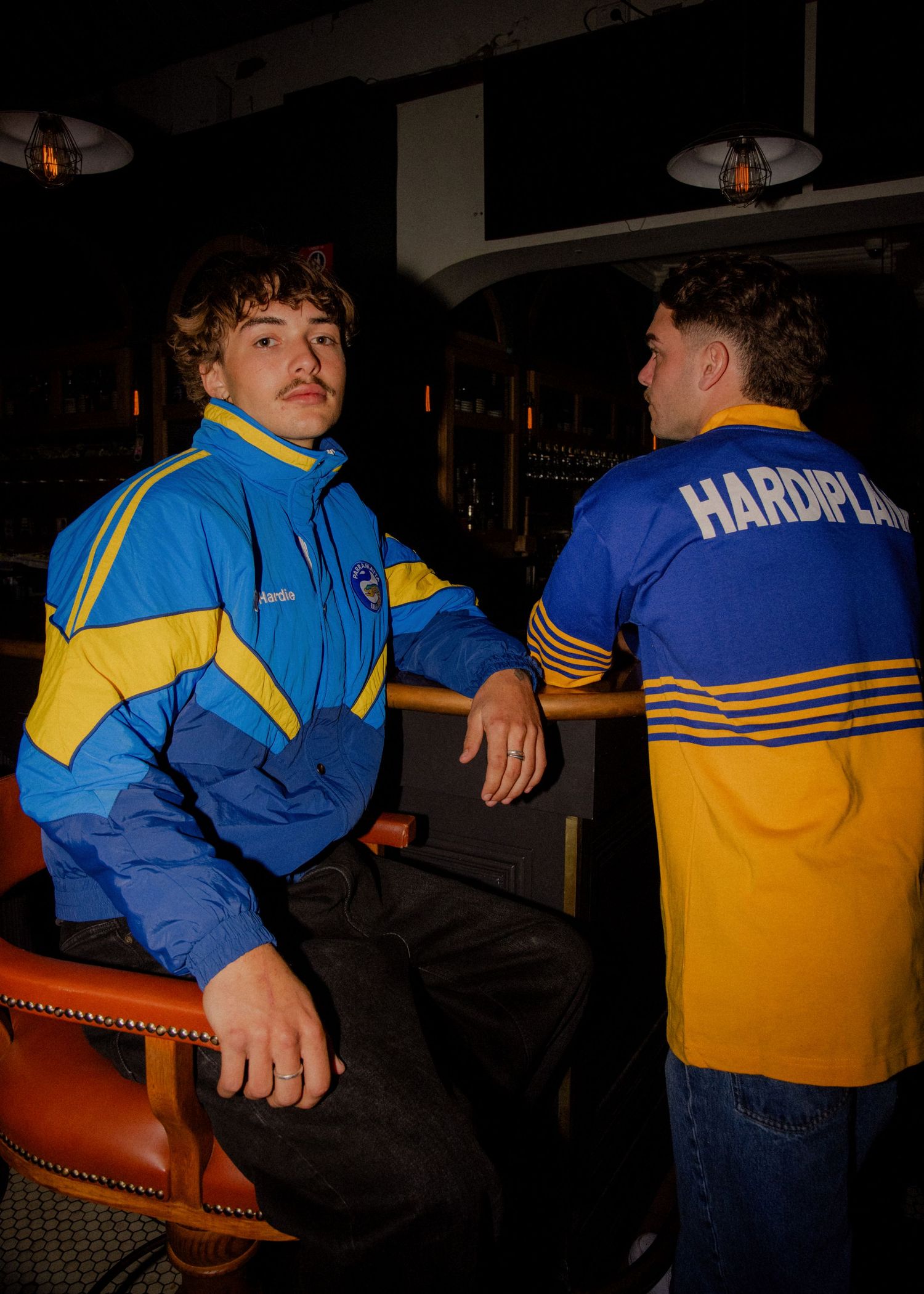 Two men wearing retro blue and yellow Parramatta Eels NRL rugby league jackets in a dark bar setting. Wollongong photographer & videographer
