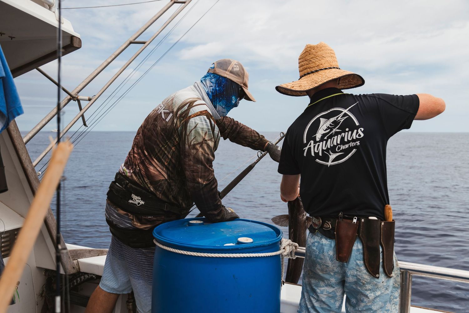 Two fishermen on a boat deck preparing equipment over a blue barrel with the ocean in the background.