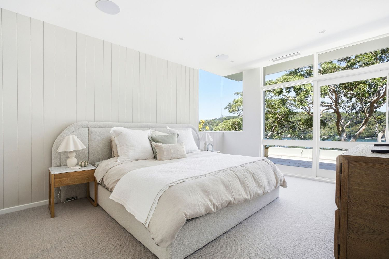 Bright master bedroom with white panelled walls, plush bedding, timber nightstand, and floor-to-ceiling windows overlooking lush trees.