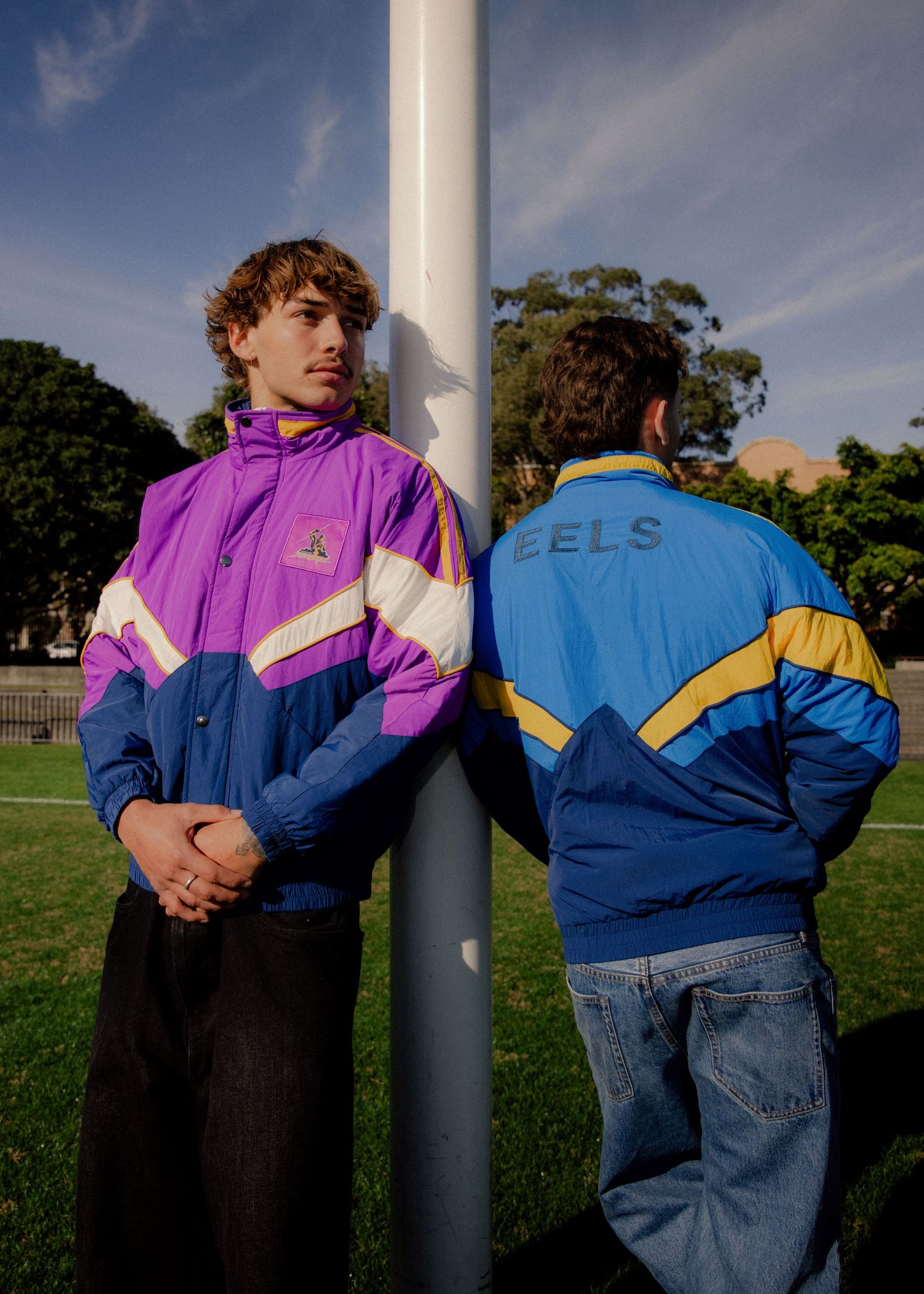 Two male models wearing retro purple and blue NRL Parramatta Eels windbreaker jackets on a sports field. Business and brand photographer
