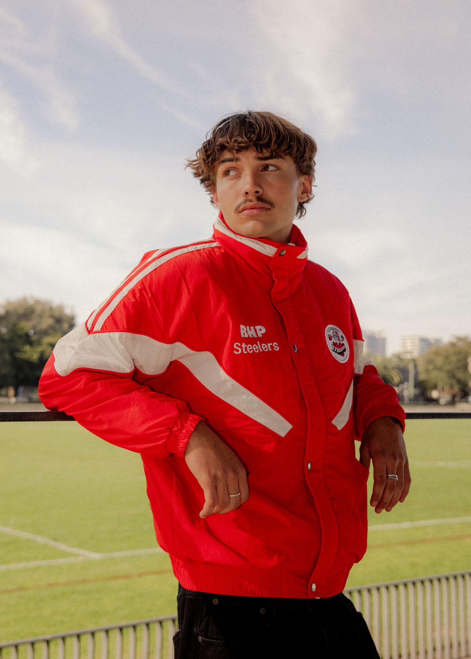 Young man in red Steelers windbreaker jacket posing outdoors near a sports field on a cloudy day.