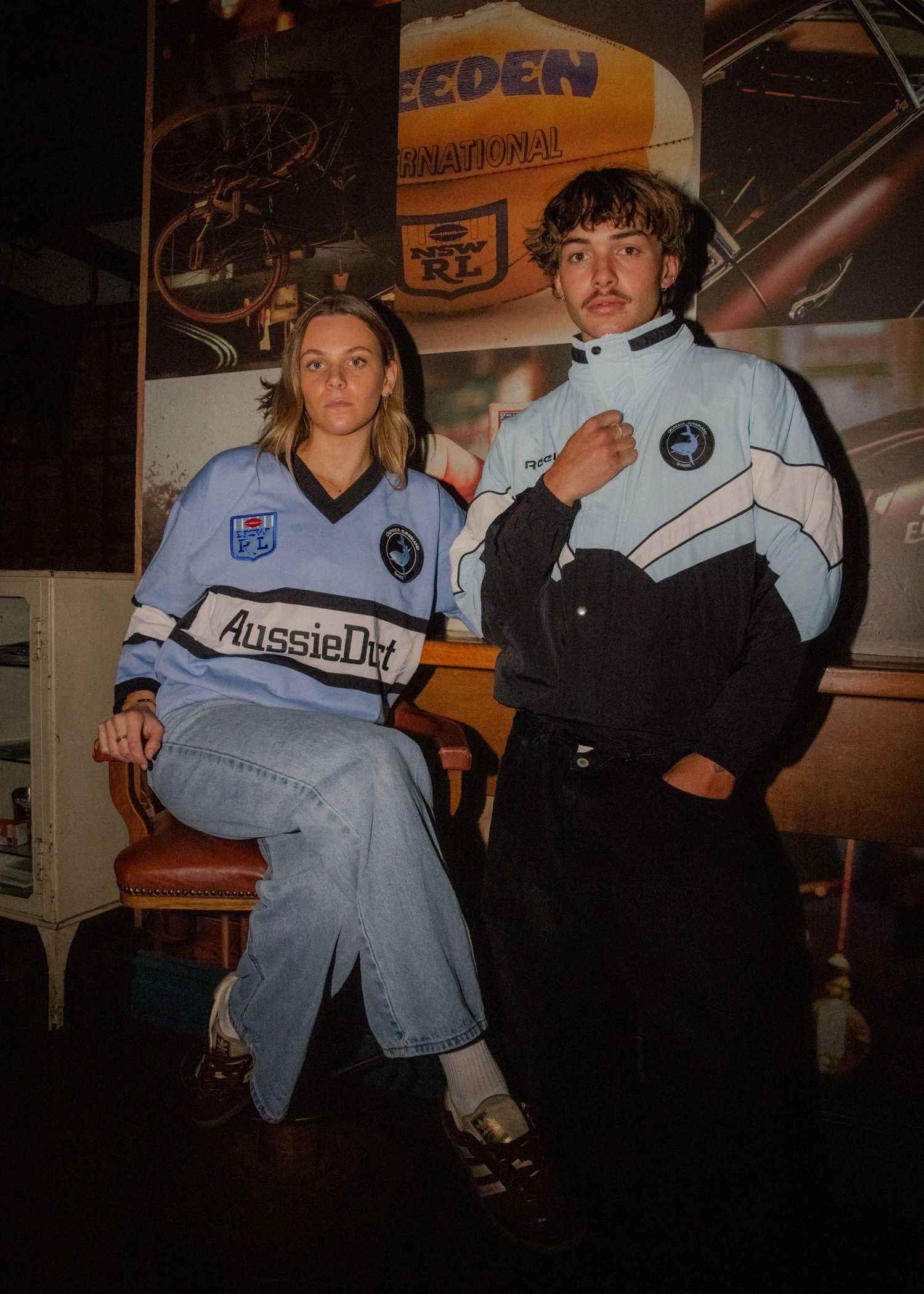Two models wearing Cronulla Sharks NRL retro jerseys and tracksuit, posing indoors against a vintage motorsport backdrop. Wollongong video