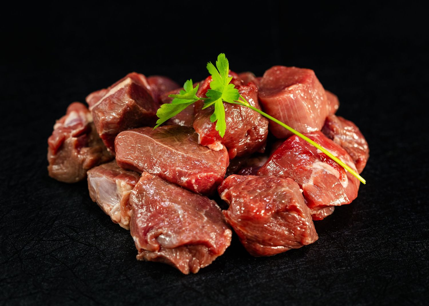 Fresh raw diced beef chunks piled on a dark surface, garnished with a sprig of fresh parsley.