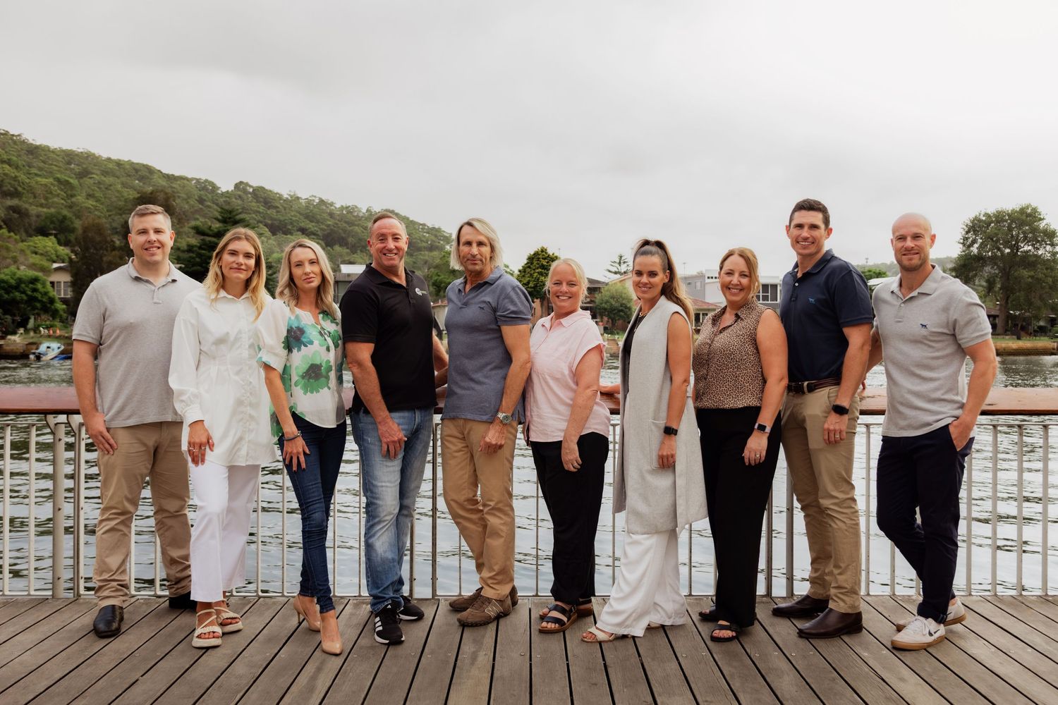 Group of ten professionals posing on a wooden deck by a river, smiling outdoors on a cloudy day.