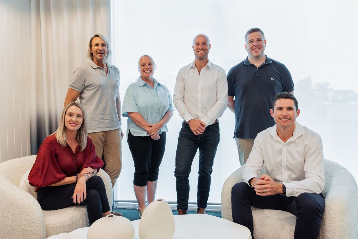 Six business professionals posing together in a modern office space with large windows and contemporary white furniture.