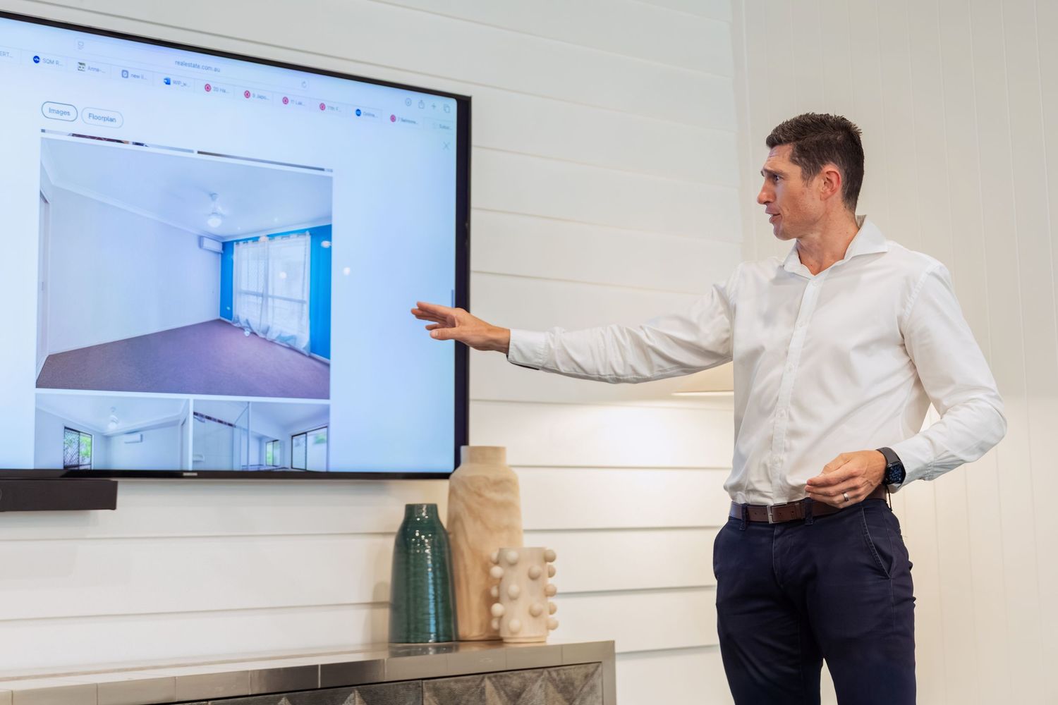 A man in a white shirt presents a room interior displayed on a large TV screen during a real estate presentation.
