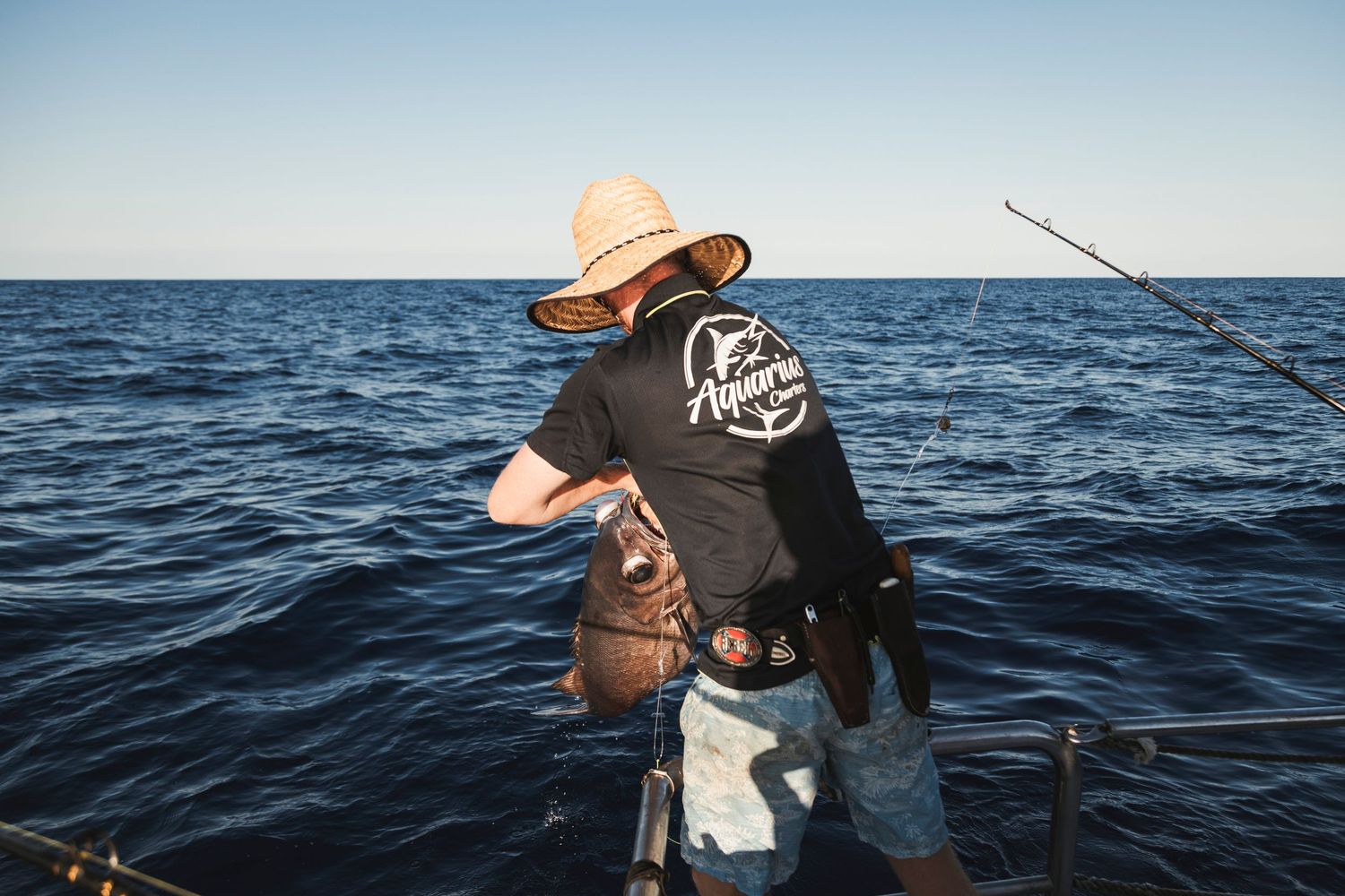 Angler on a boat holding a caught fish over the ocean, wearing a straw hat and black fishing shirt.