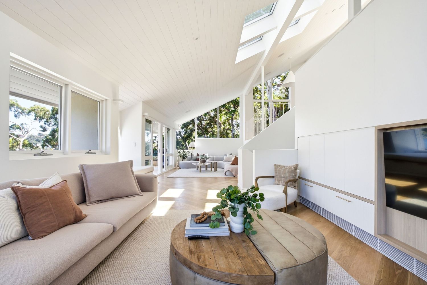 Bright modern living room with vaulted white ceiling, beige sofa, round wood coffee table, and open view to outdoor patio.
