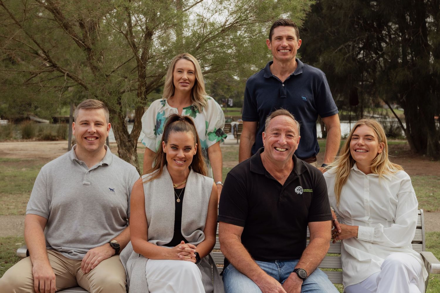 Group of six smiling adults posing outdoors in a park setting, three seated and three standing behind them.