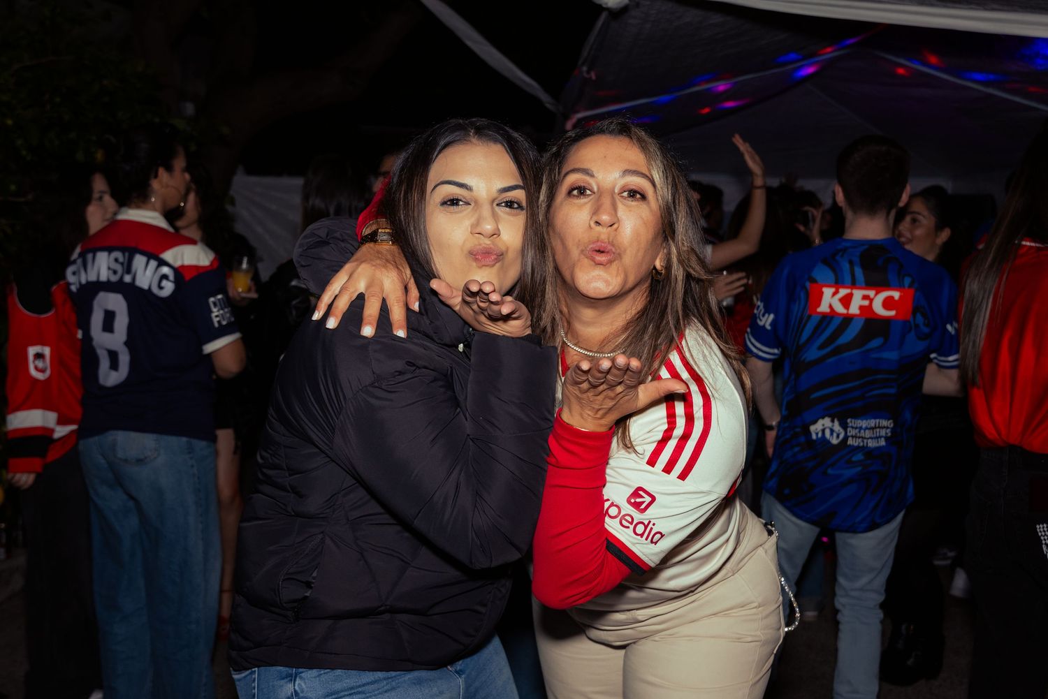 Two women making kissy faces at a sports fan event, wearing jerseys and casual jackets outdoors at night.