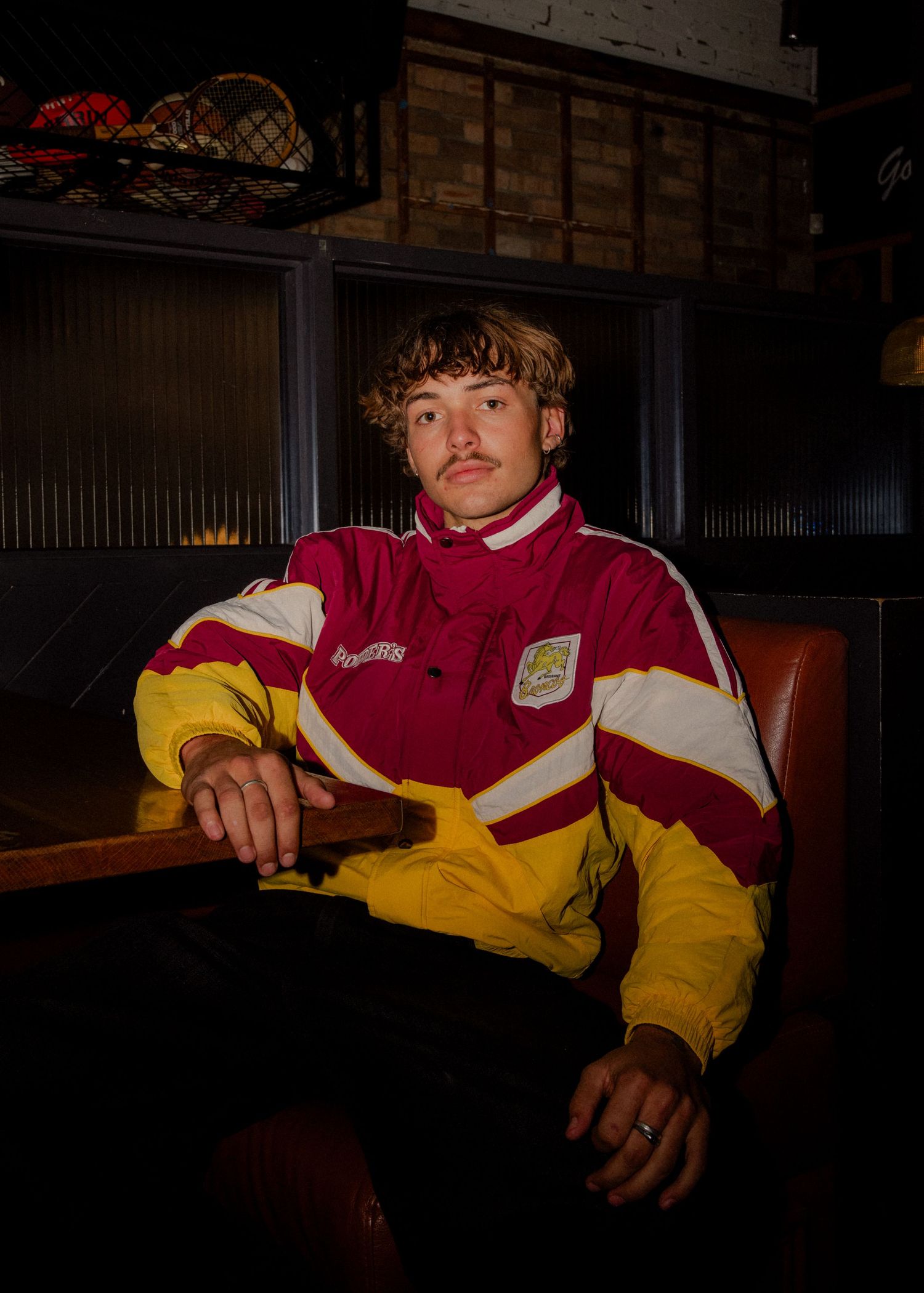 Young man wearing retro red and yellow Aston Villa tracksuit jacket seated in dimly lit bar booth. Business and brand photographer sydney