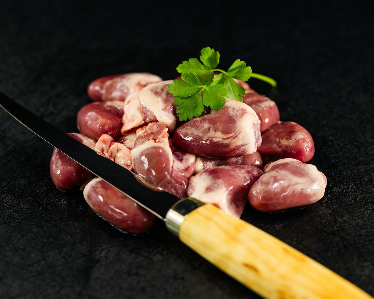 Food and product photography and videography Raw chicken hearts with parsley garnish and wooden knife on black surface.