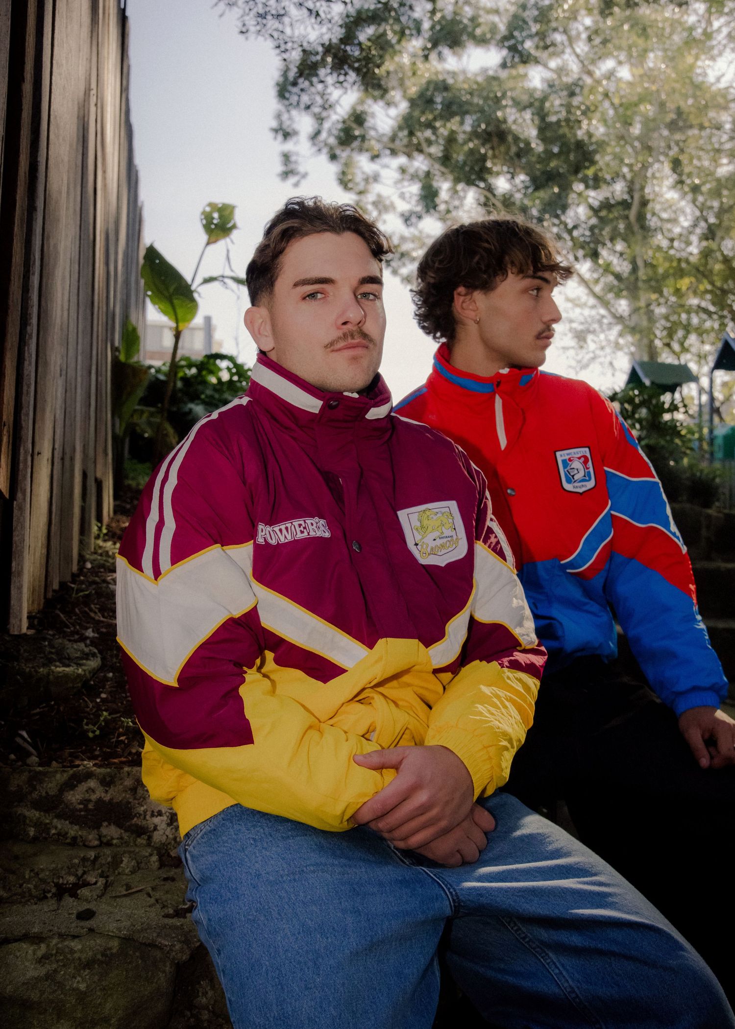 Two men wearing vintage NRL maroon and blue puffer jackets sitting outdoors in natural light.