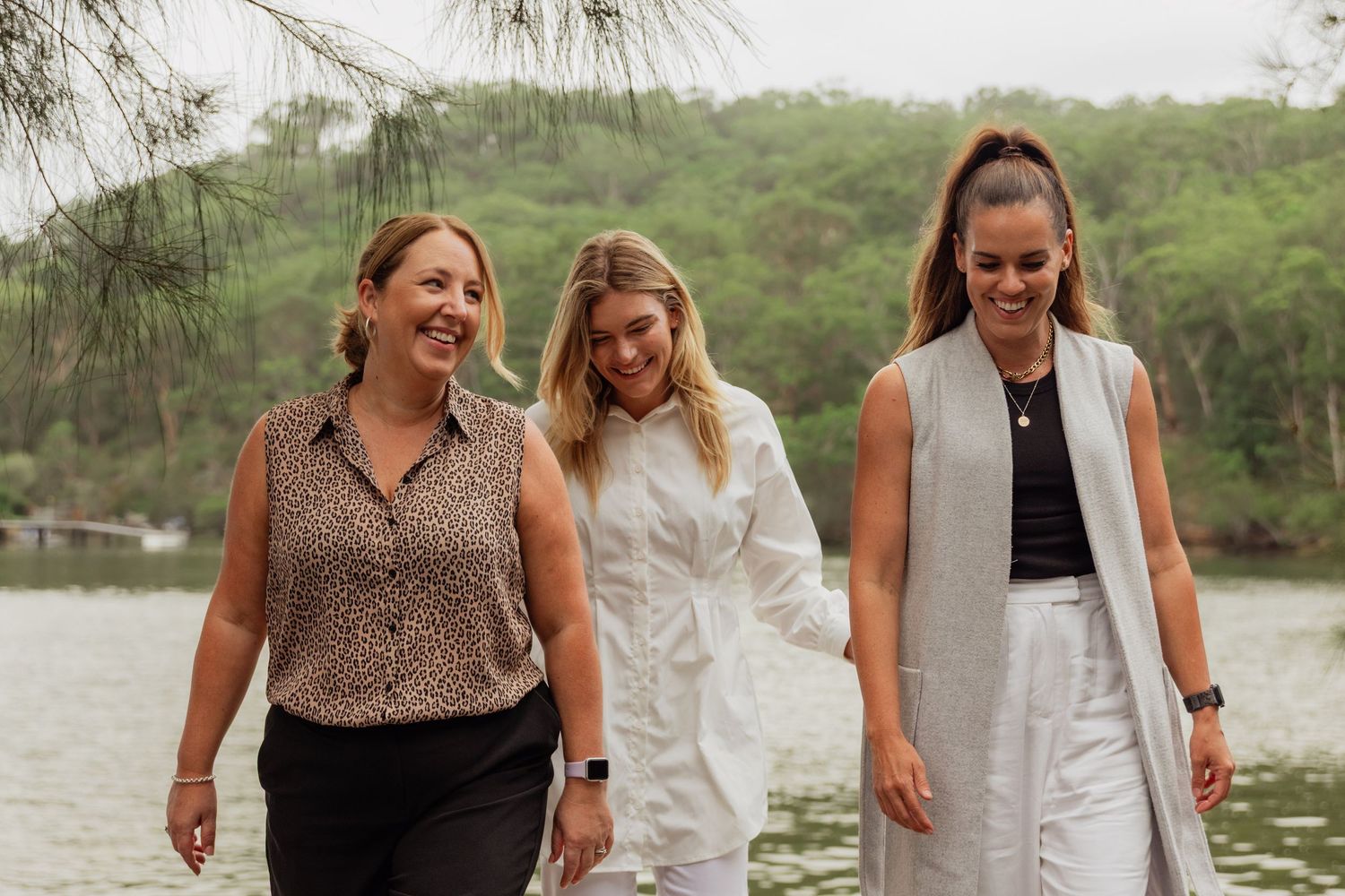 Three women smiling and walking together outdoors near a lake with lush green trees in the background.