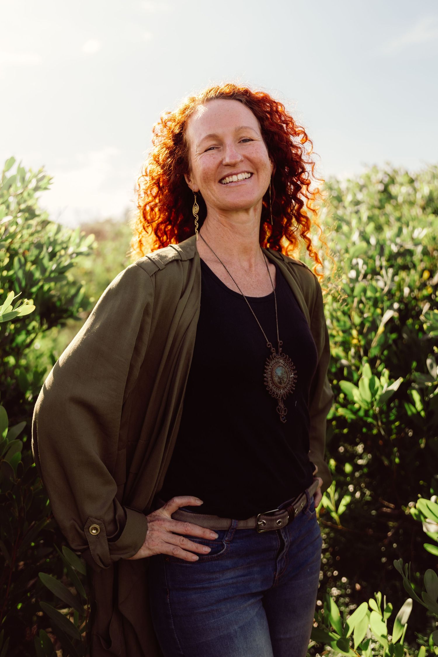 Smiling woman with curly red hair wearing green cardigan and jeans standing outdoors among lush green plants.
