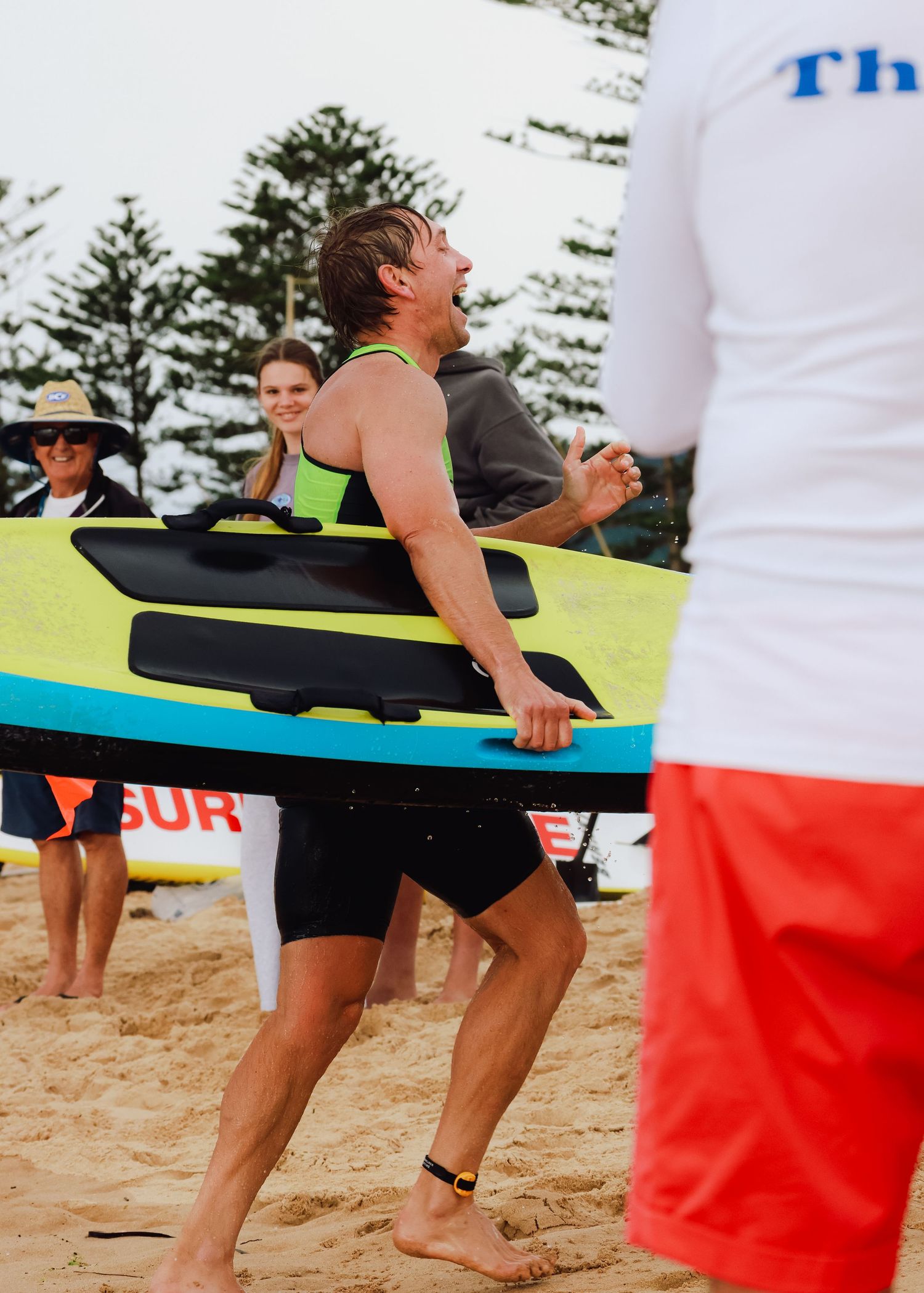 Man laughing on sandy beach holding colorful surfboard during surf competition with spectators in background.