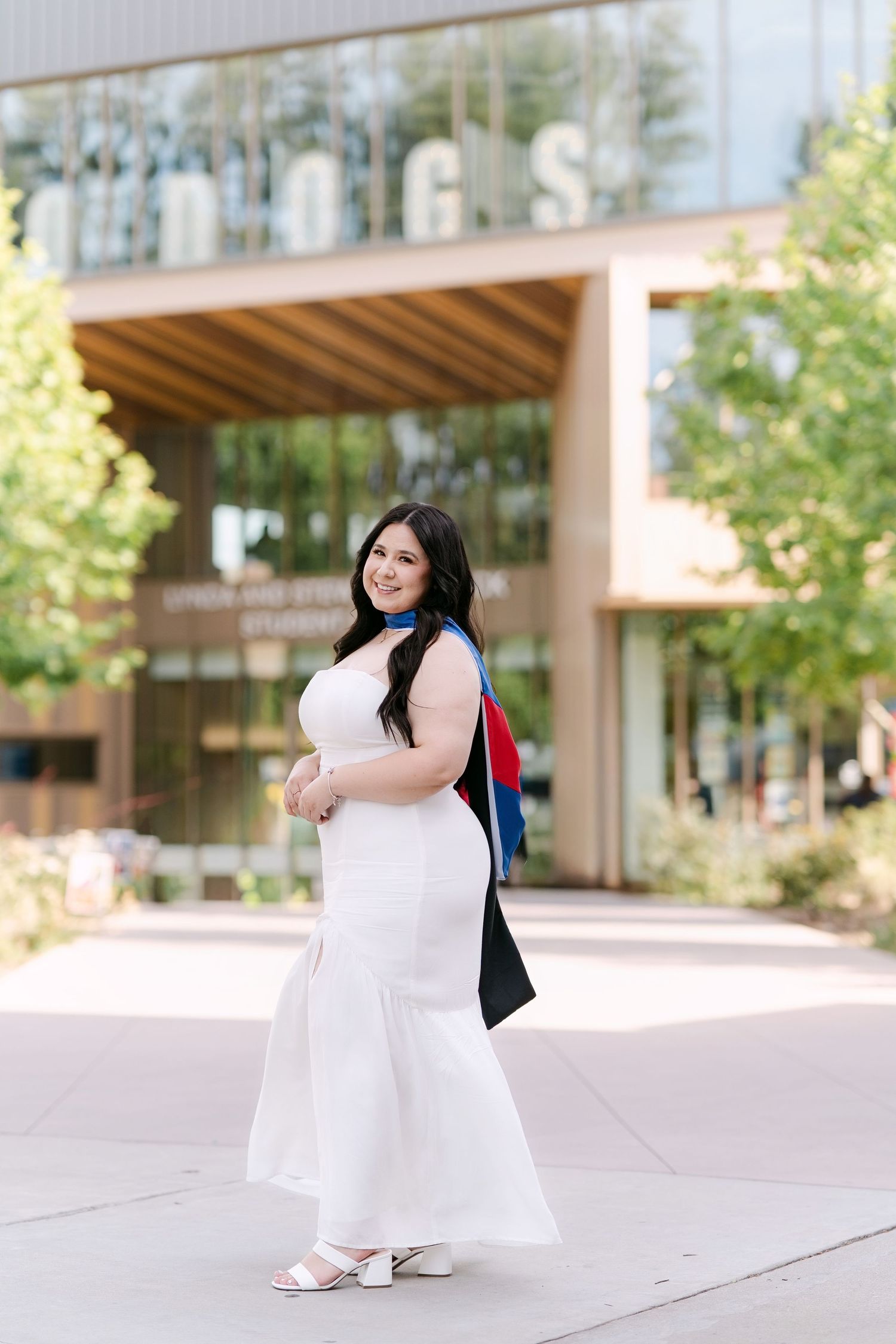 A young woman in a white mermaid dress and graduation regalia poses outdoors in front of a modern university building.