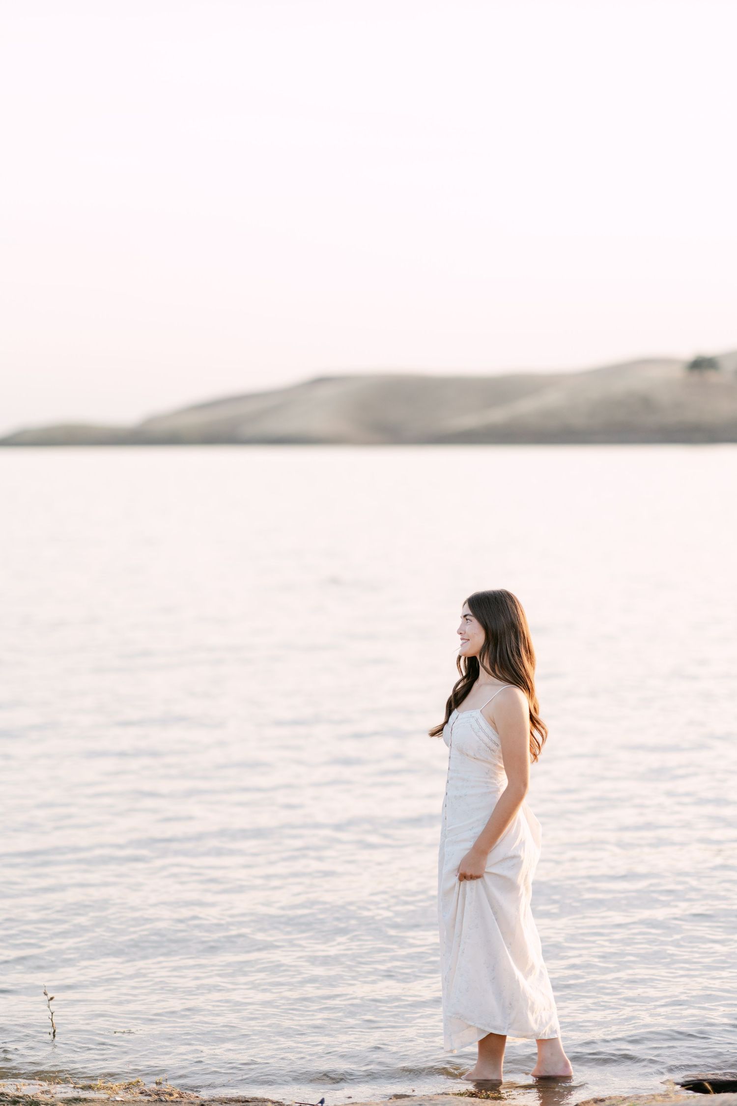Young woman in white dress standing by calm water at sunset with sandy hills in the background.