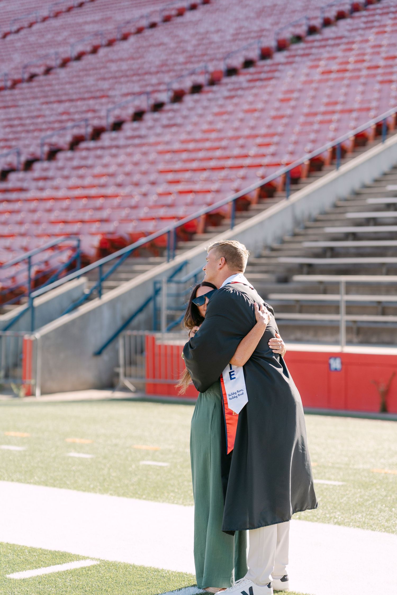 Two people embracing on a football field with empty red stadium seats in the background.