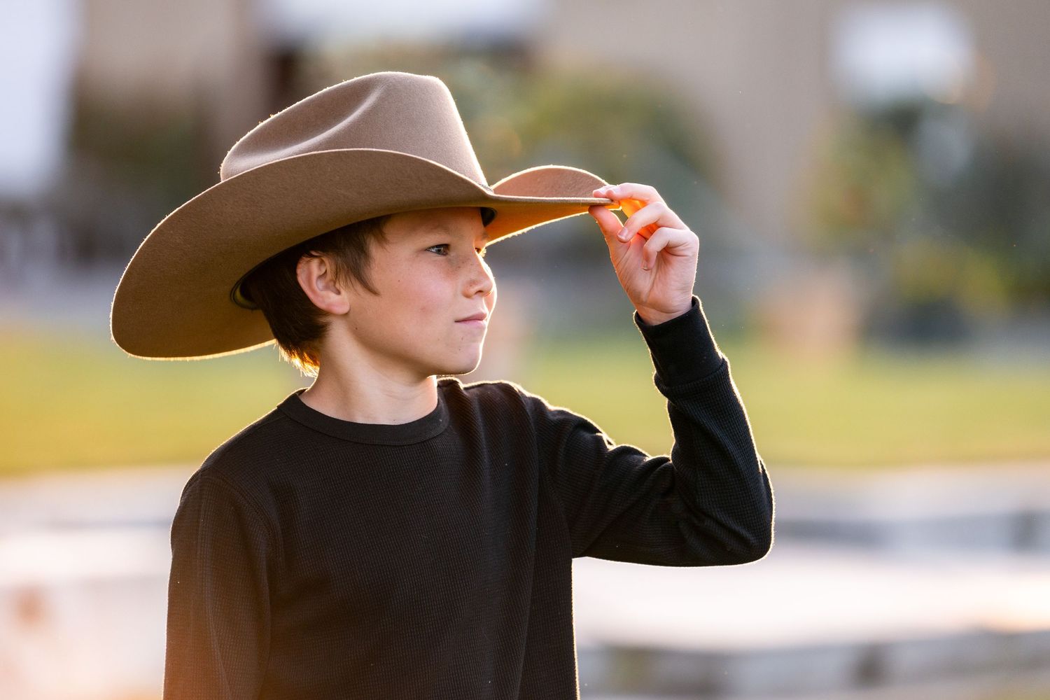 A person in a black sweater and brown cowboy hat tips their brim in the evening light.