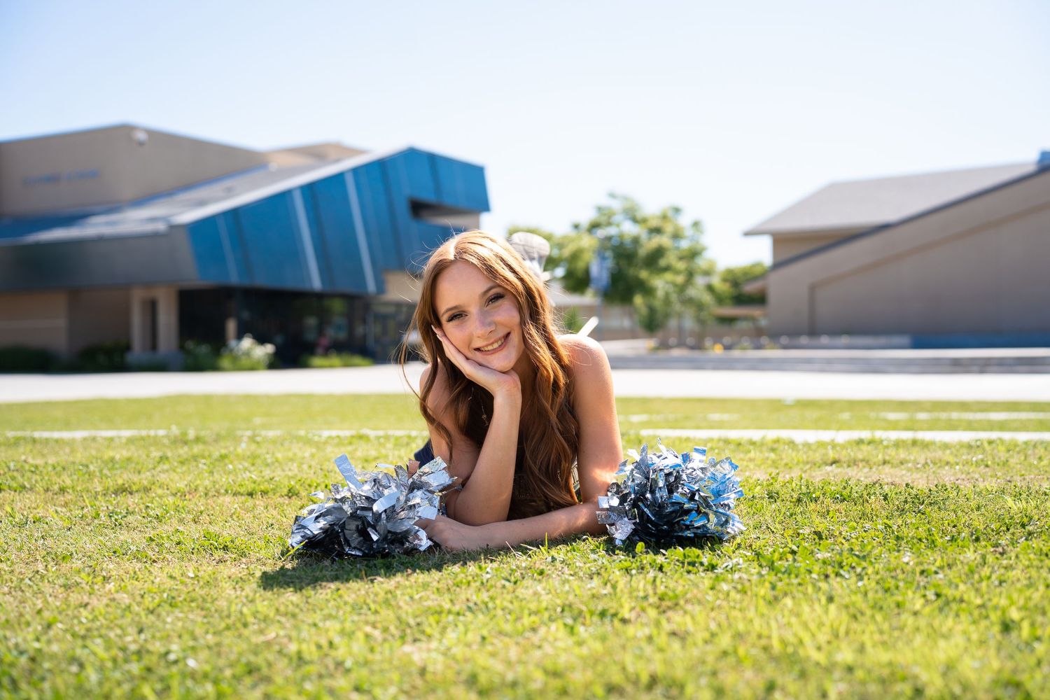 Cheerleader lies on grass field with pom poms during outdoor photo session.