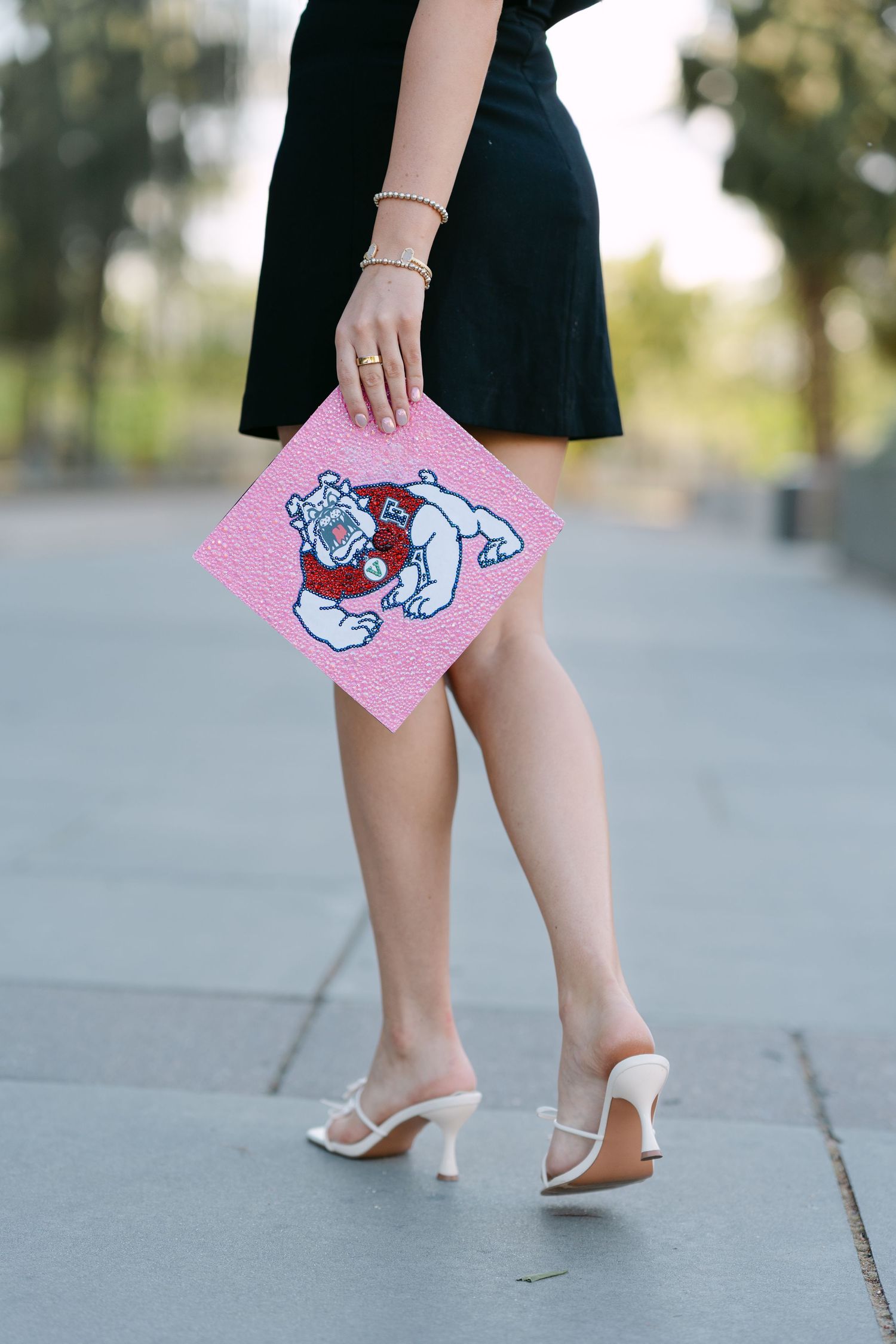 Graduate holding a pink Fresno State Bulldogs decorated graduation cap while wearing a black dress and white heels.