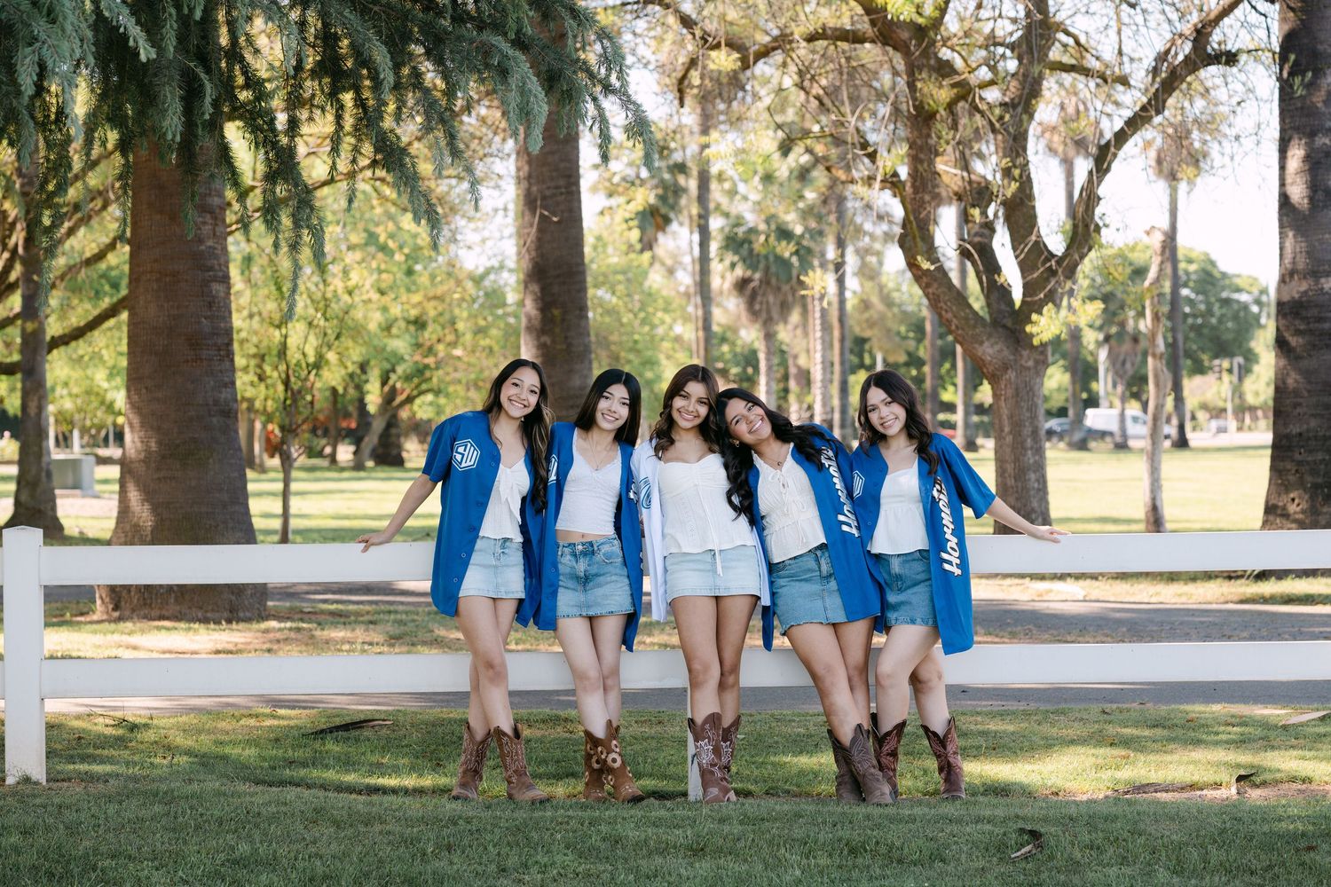 Six young women in blue jackets and denim shorts pose together outdoors in a tree-lined park setting.
