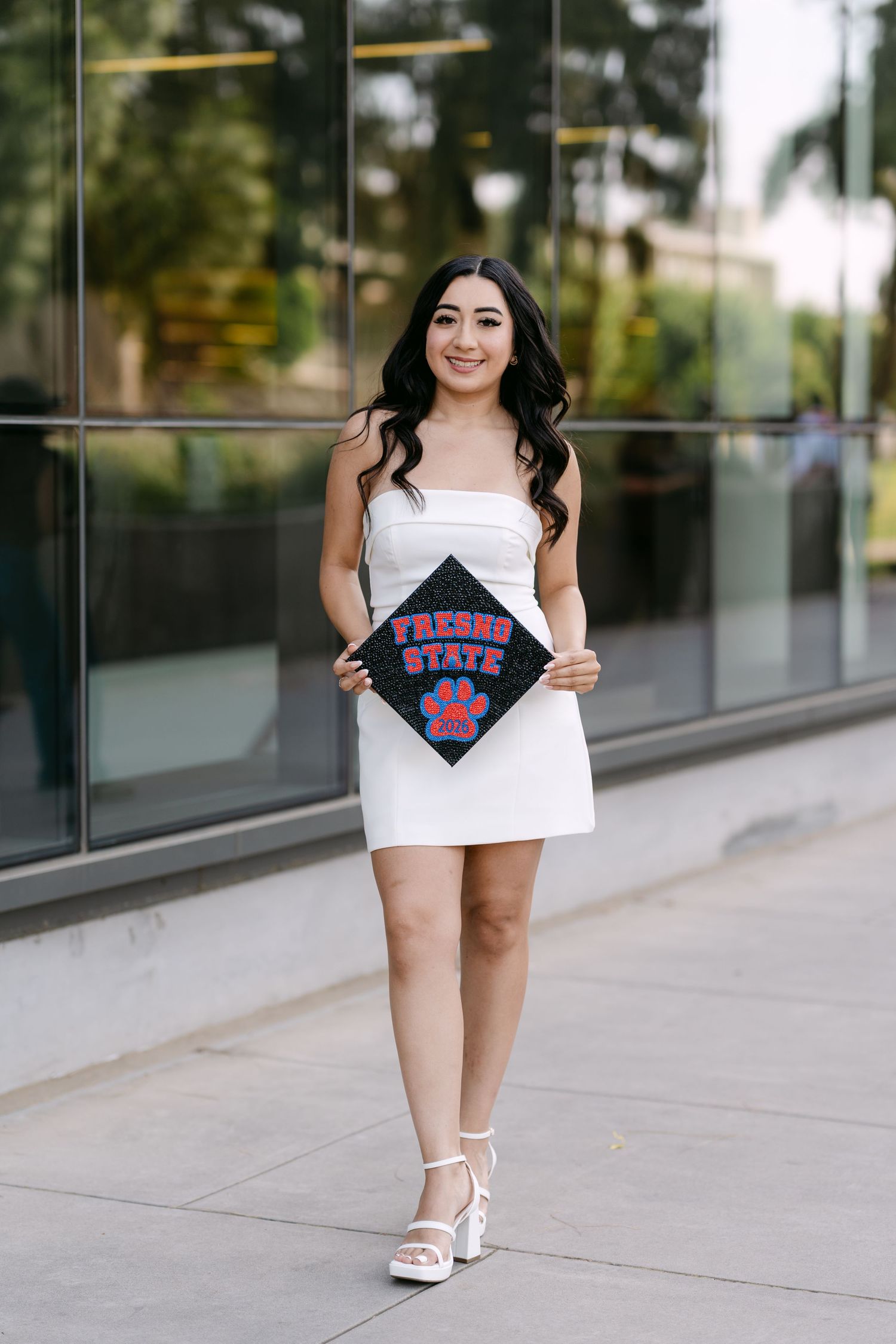 Young woman in white dress holding Fresno State graduation cap, posing outdoors for graduation photos.