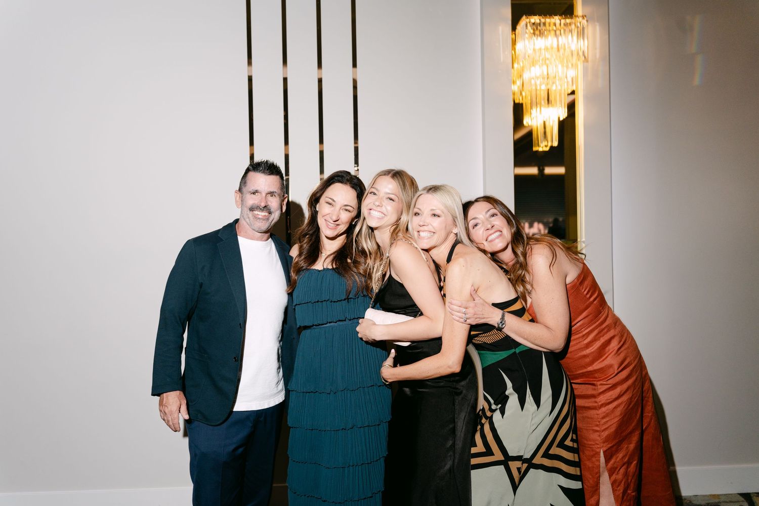 Group of five friends smiling and hugging at an elegant indoor event, dressed in formal attire.