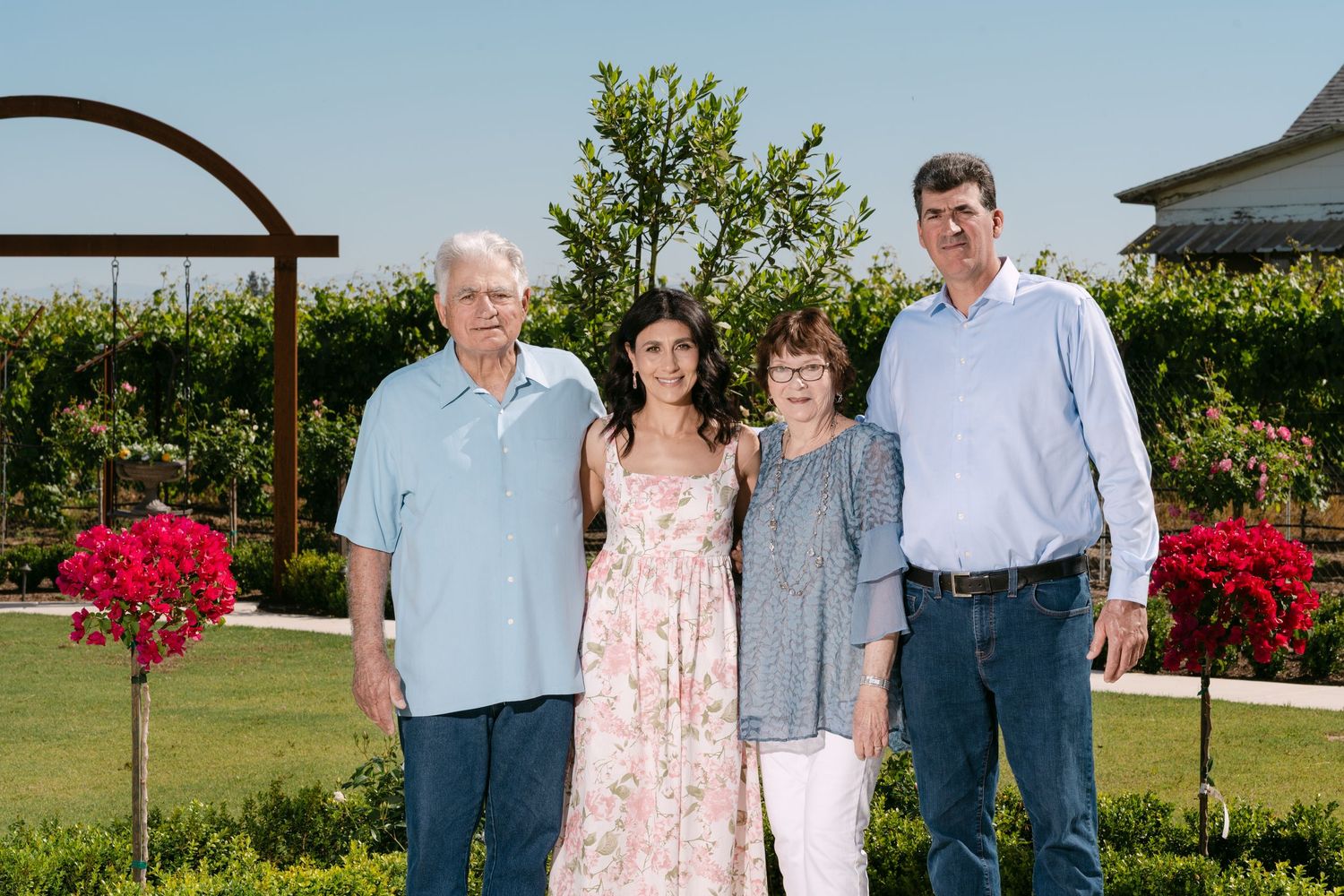 Family of four posing outdoors in a garden with red flowers and green trees on a sunny day.
