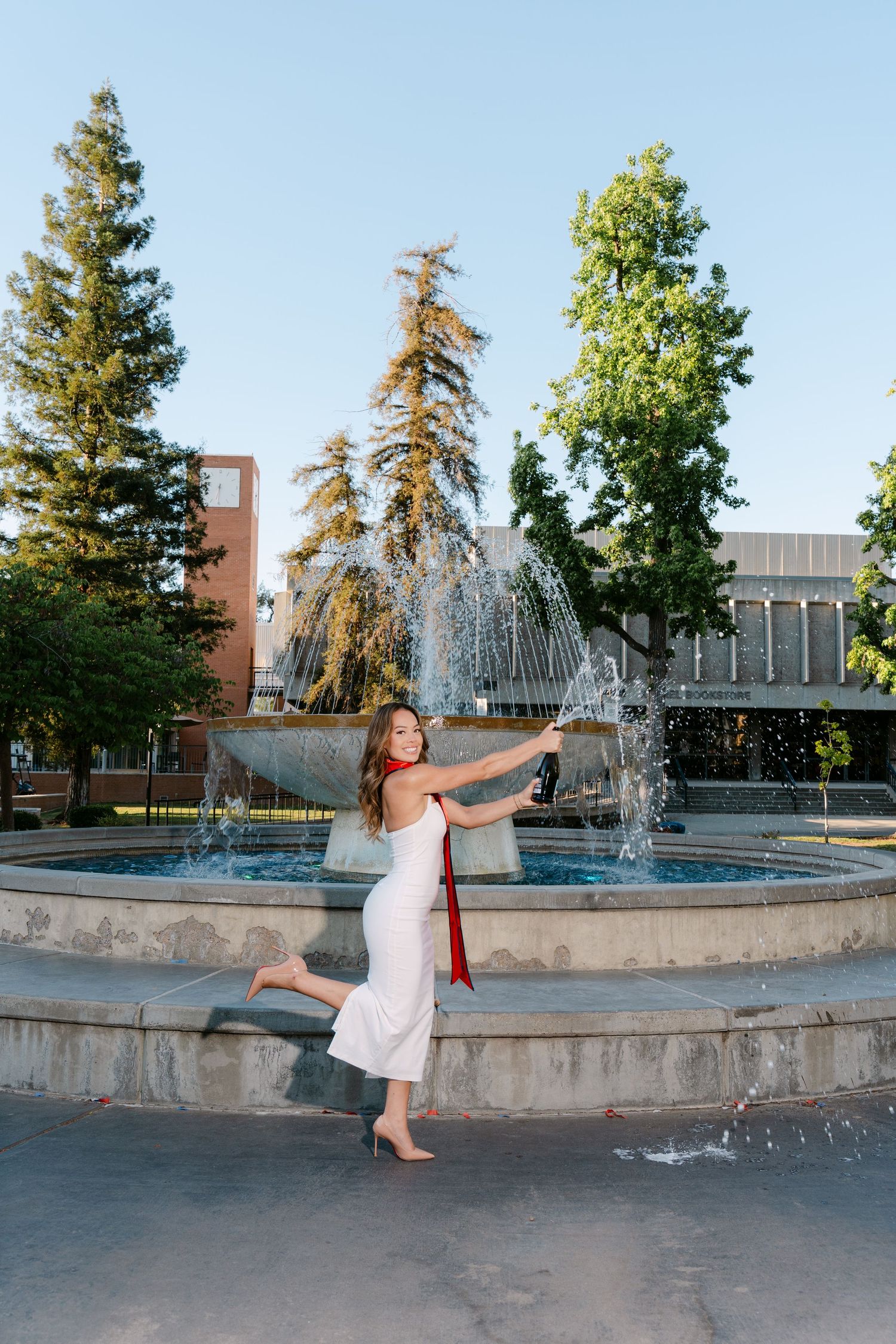 Young woman in white dress and red sash poses gracefully by a campus fountain with trees in the background.