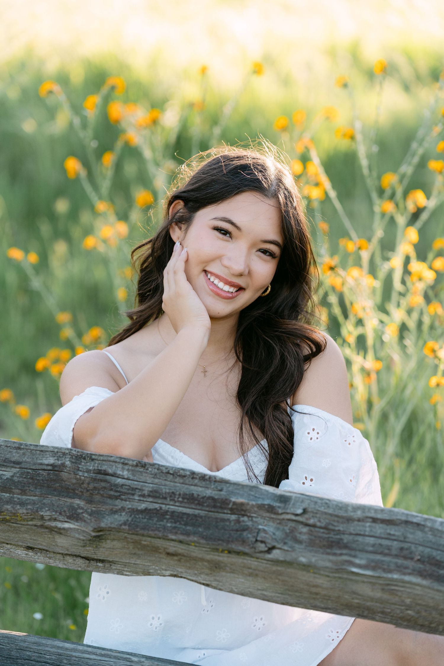 Smiling young woman in white off-shoulder dress leaning on wooden fence with yellow wildflowers in background.