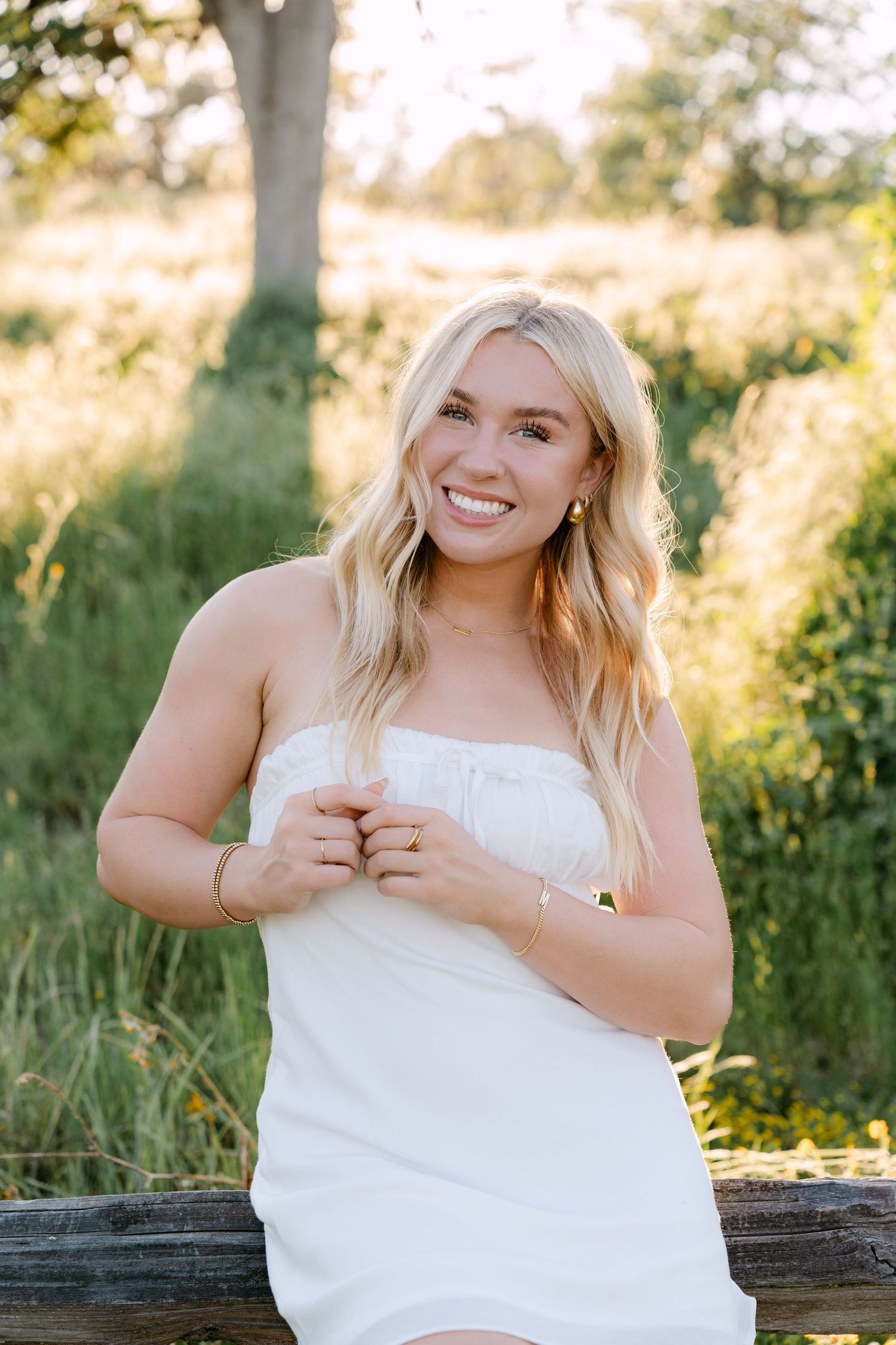 Smiling blonde woman in white strapless dress sitting on wooden fence in sunny outdoor field.