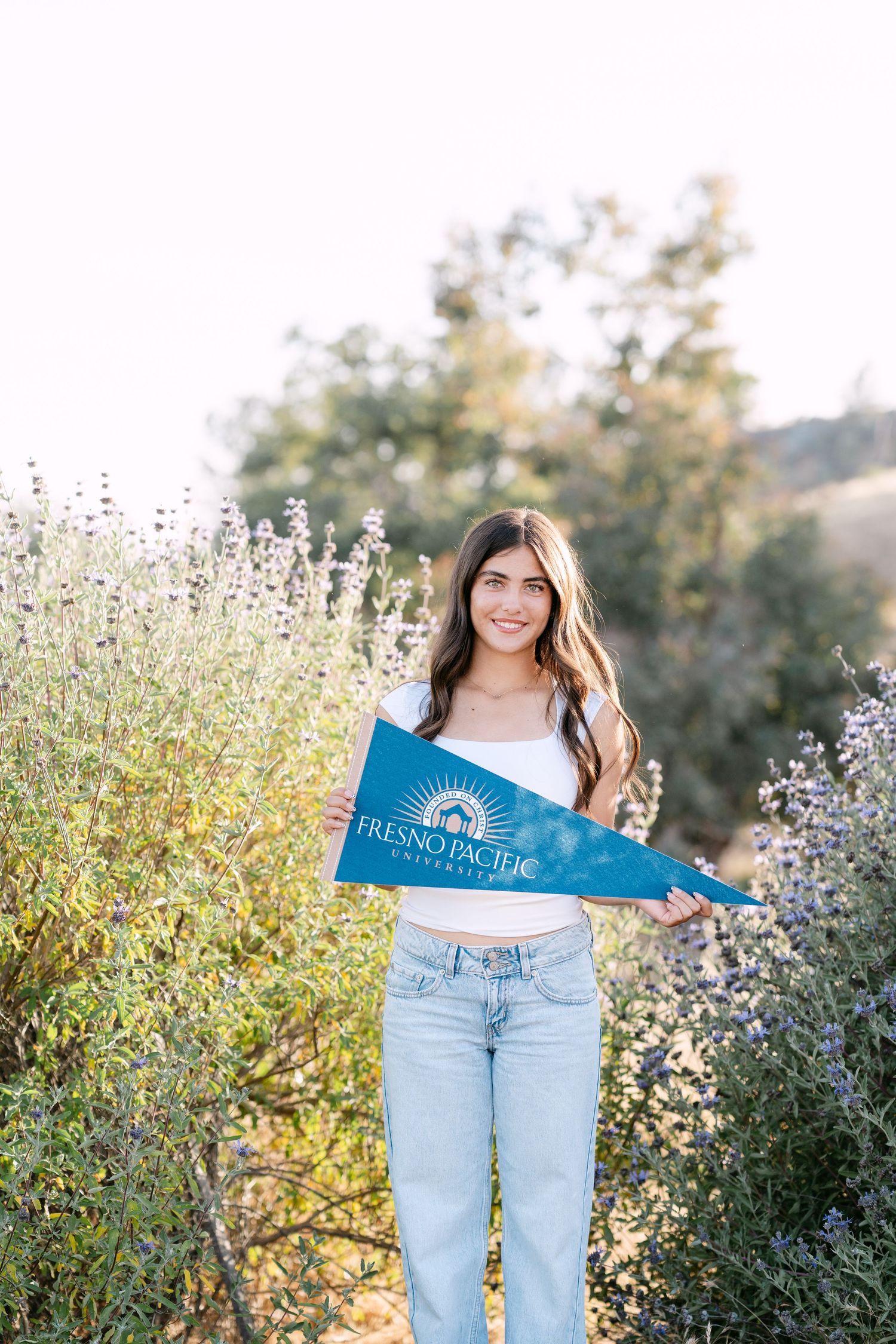 Young woman smiling outdoors holding a Fresno Pacific University pennant flag surrounded by blooming wildflowers.