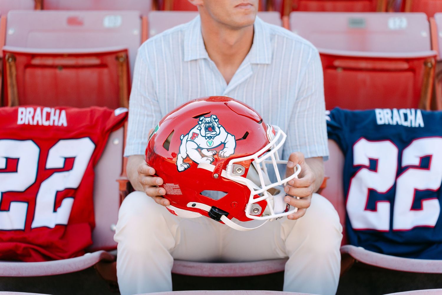 Person holding a red Fresno State Bulldogs football helmet in stadium seats with jerseys numbered 22.
