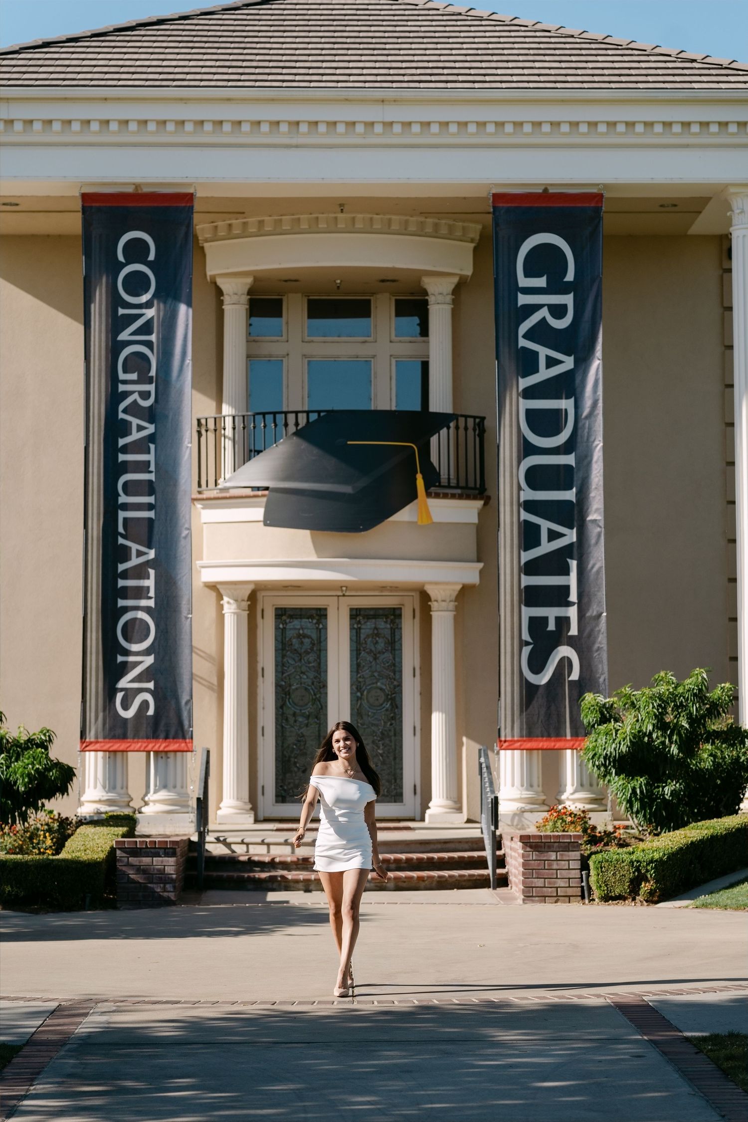 Young woman in white dress poses before mansion with Congratulations Graduates banners and graduation cap decoration.