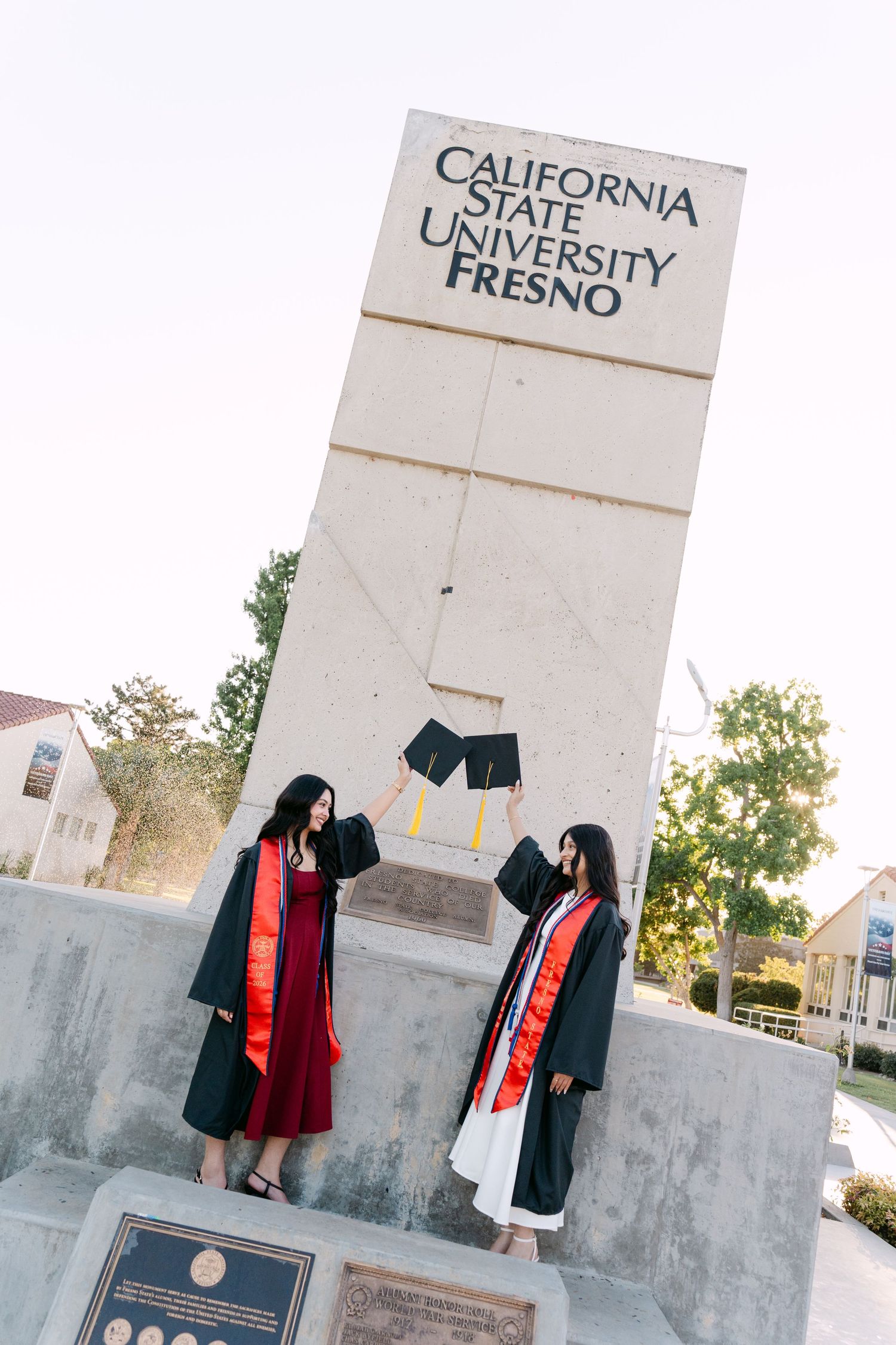 Two graduates in black robes hold caps at California State University Fresno monument sign.