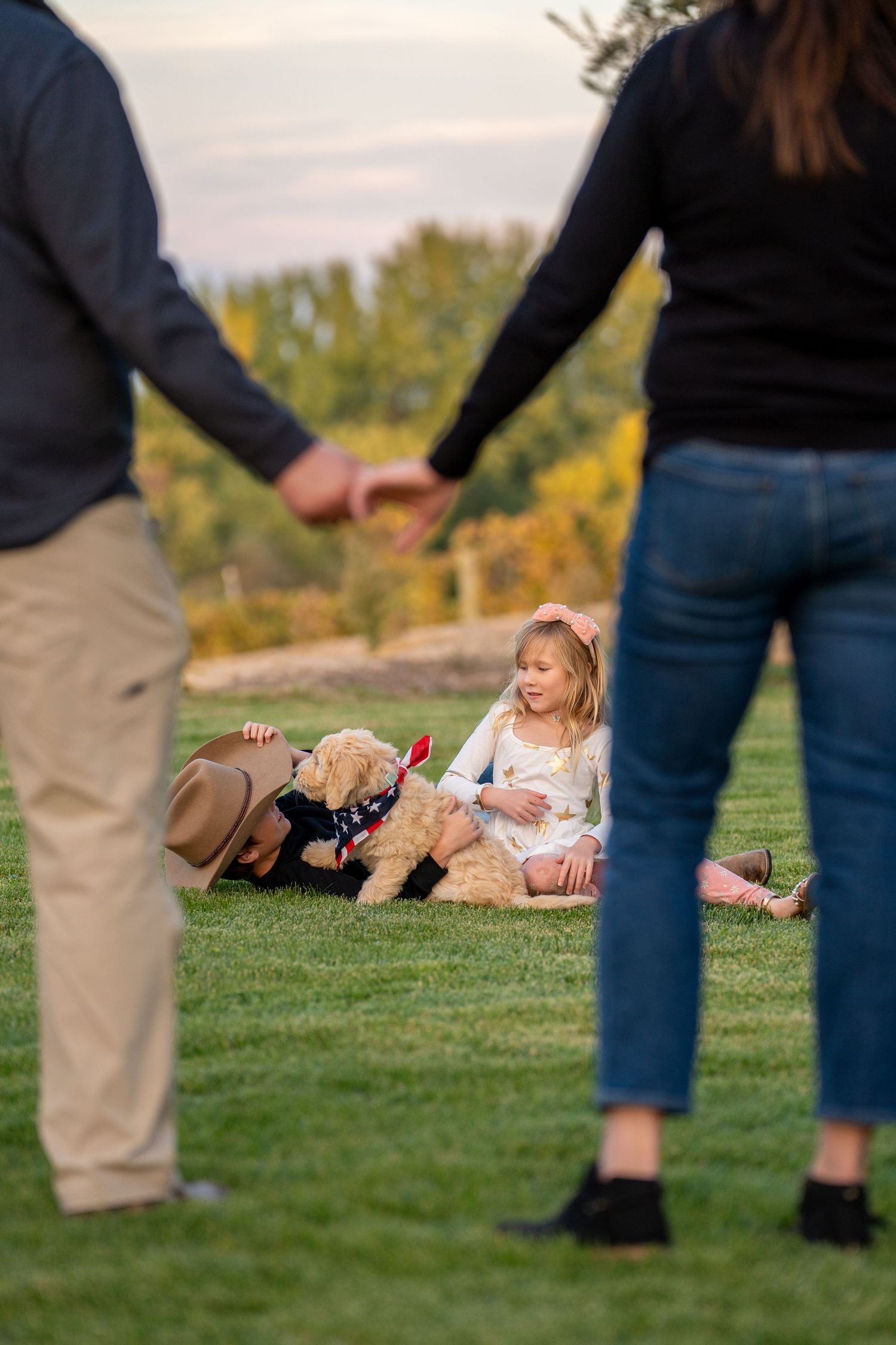 A candid family moment with a dog and child playing on grass while parents hold hands.