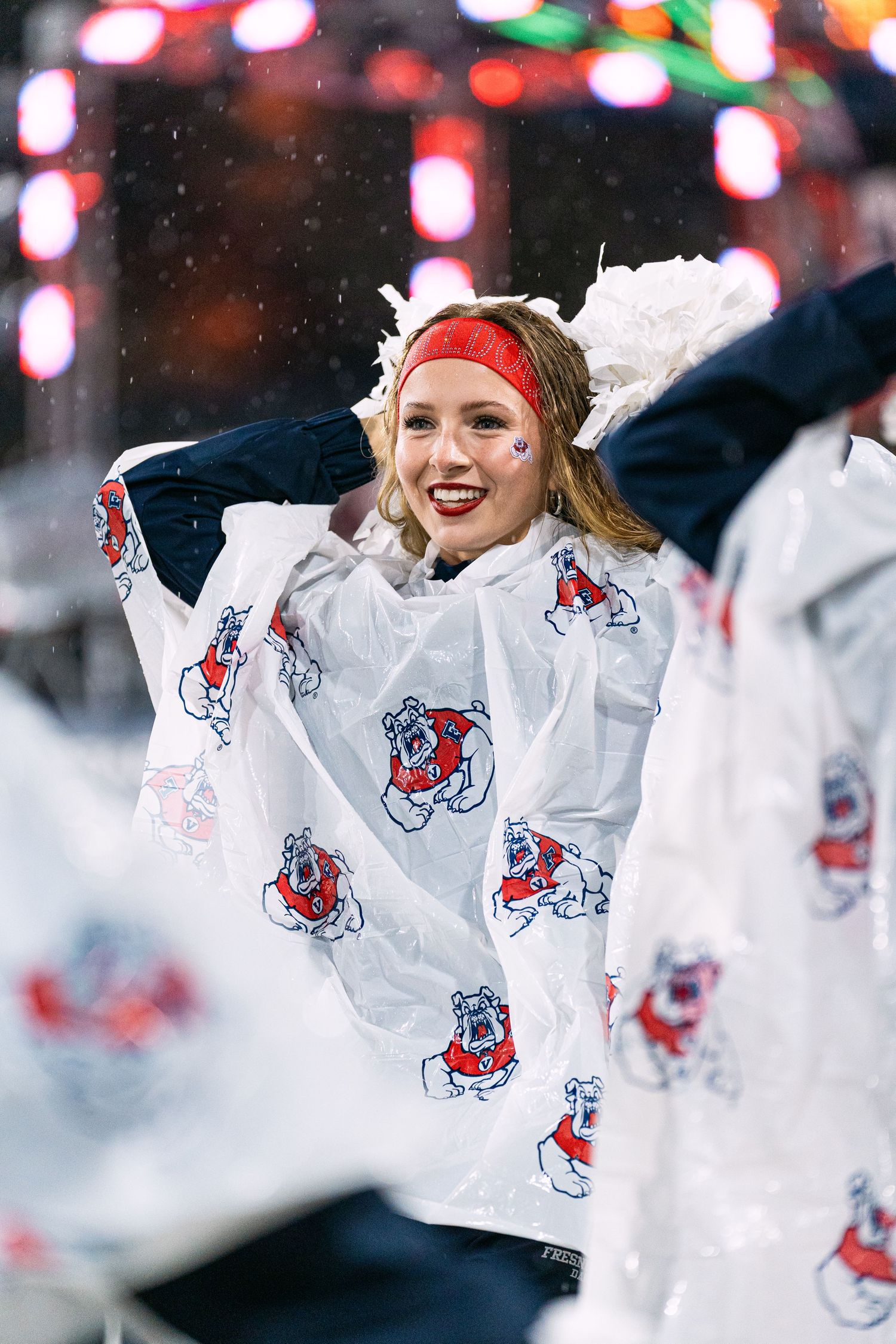 Cheerleaders in white jackets with team logos perform during a snowy sports event with colorful lights in background.