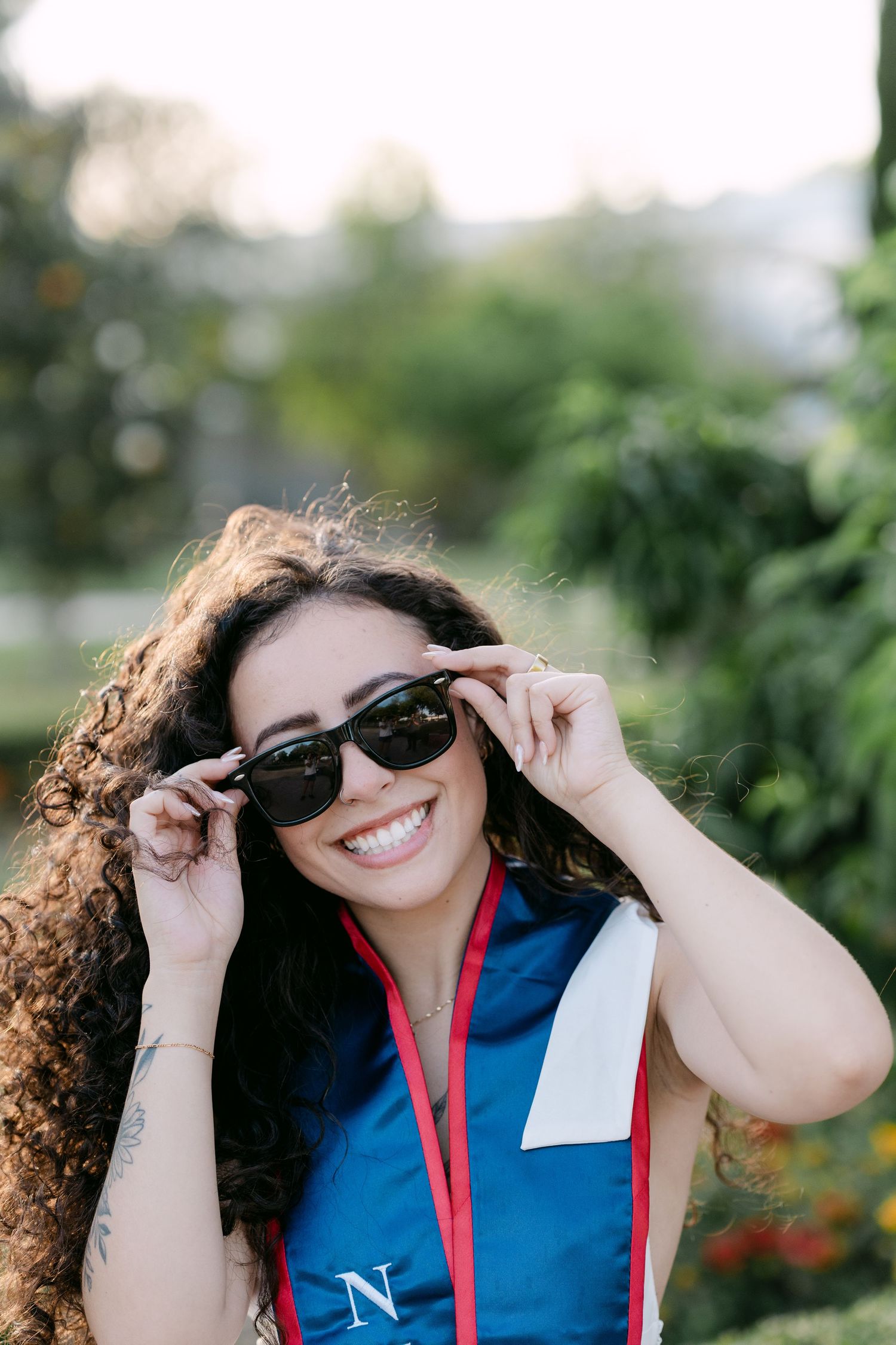 Smiling nursing graduate with curly hair adjusting sunglasses outdoors in blue graduation sash.