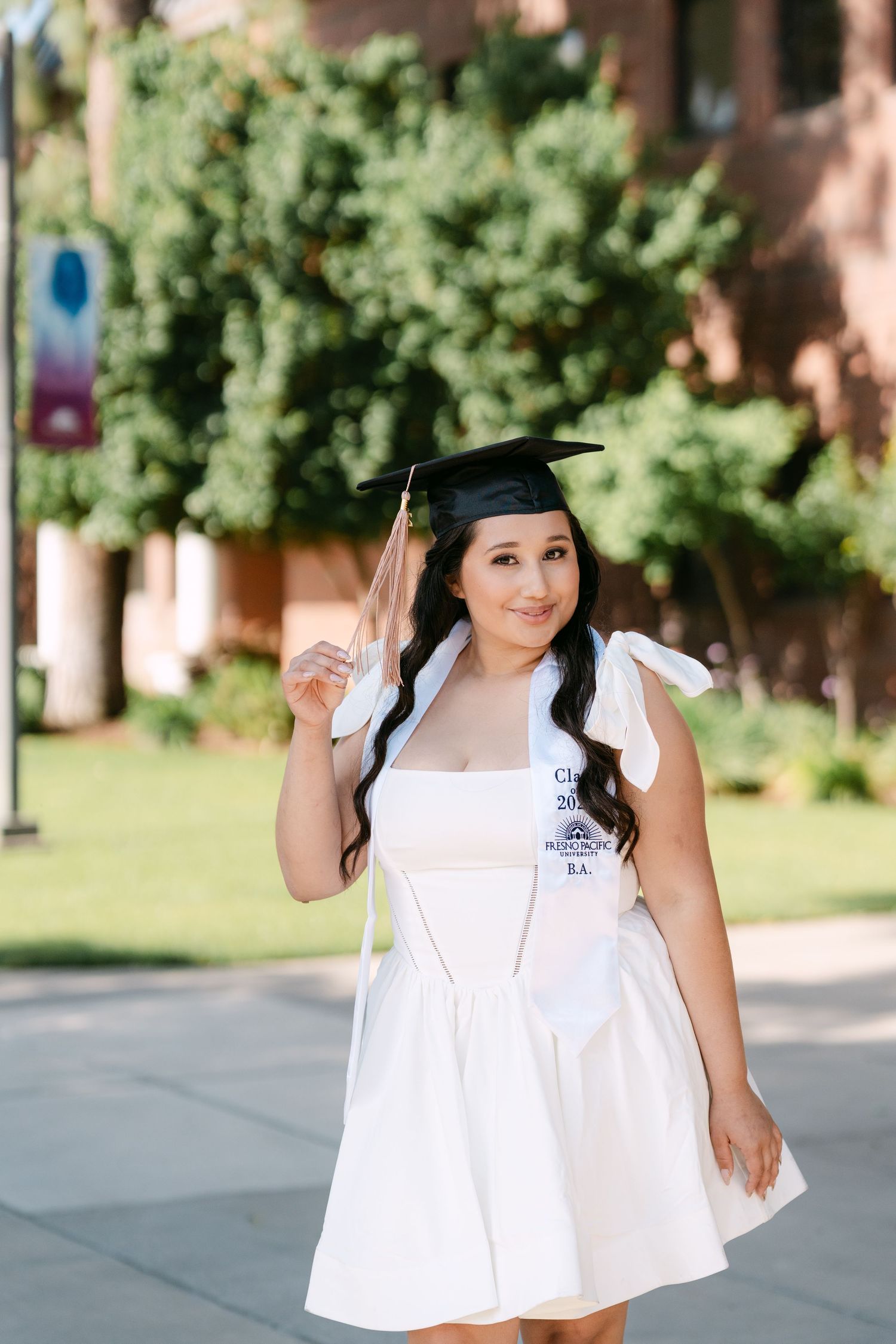 Young woman in white dress and graduation cap poses outdoors on campus, holding tassel during graduation photo session.