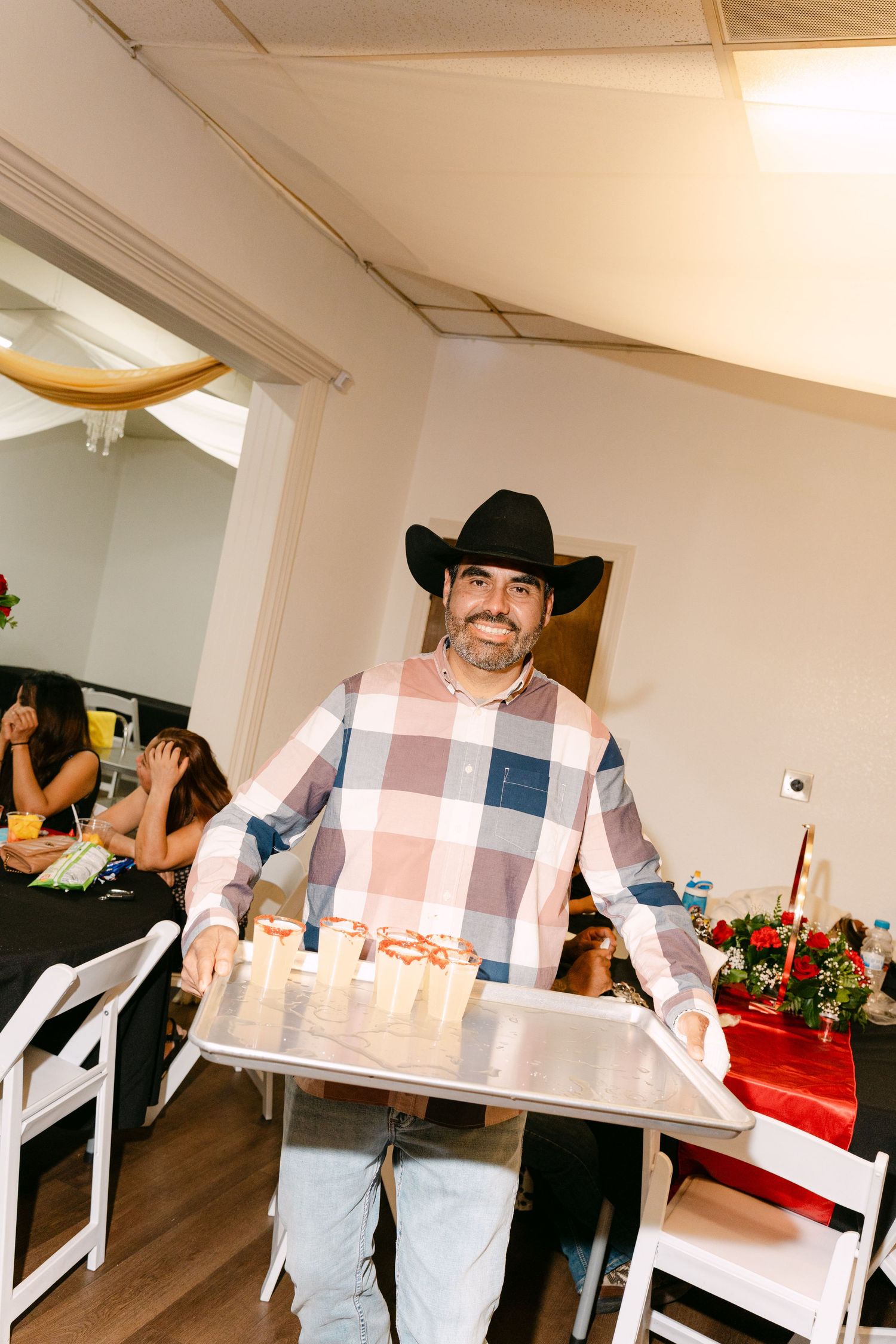 Person wearing a plaid shirt and black hat smiles while holding a large white table or surface.