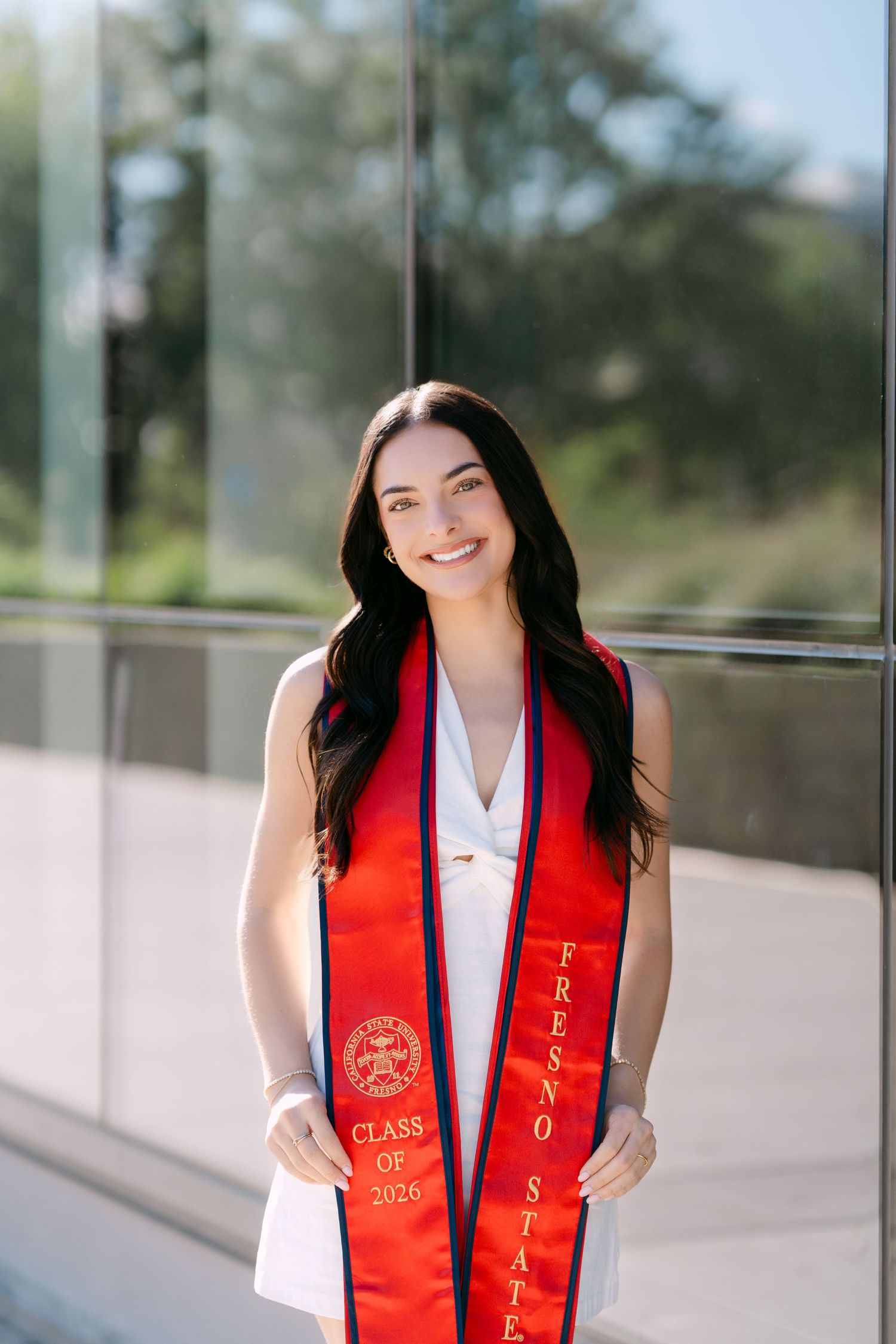 Smiling young woman wearing a red Fresno State Class of 2026 graduation stole over a white dress outdoors.