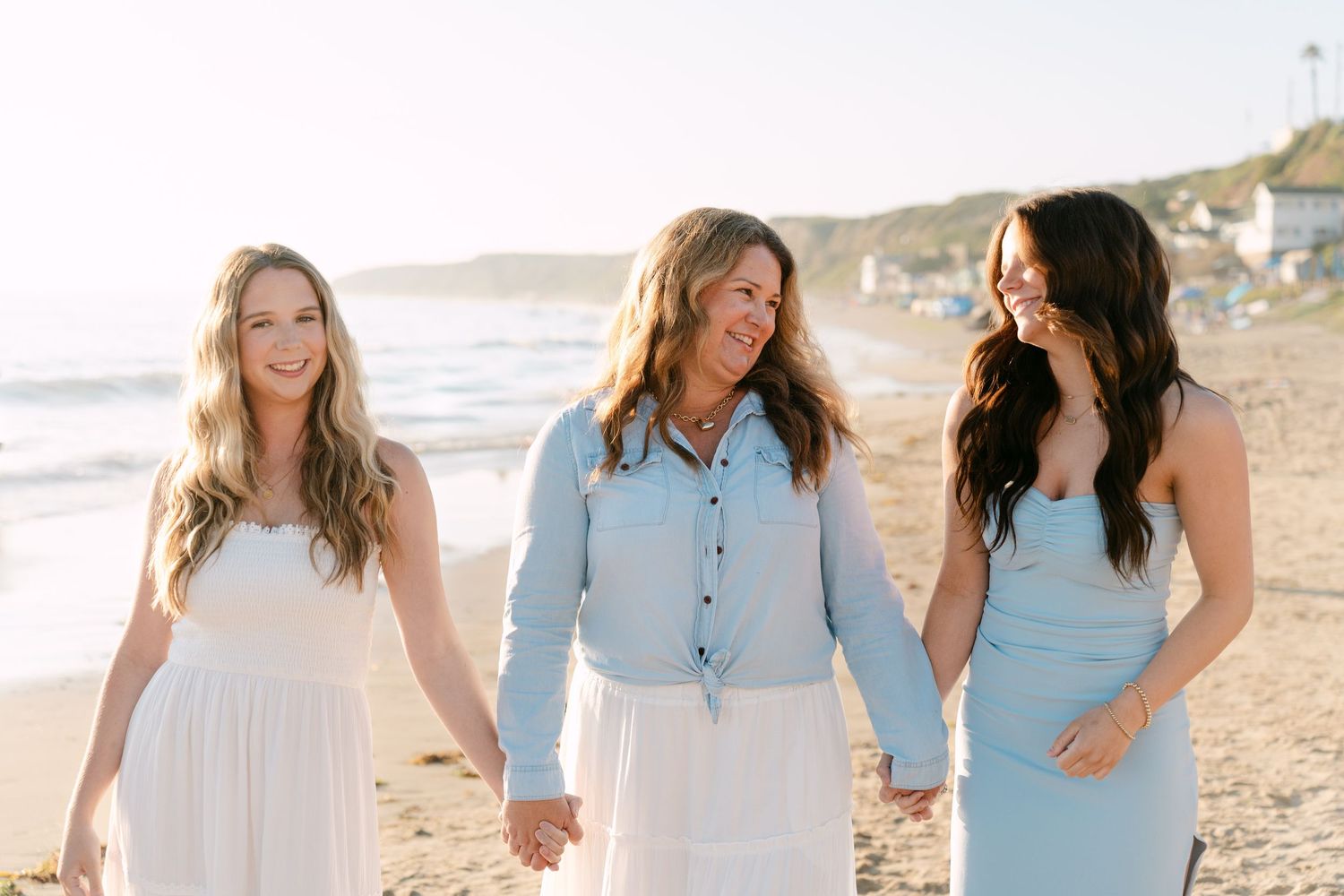 Three women holding hands and smiling on a sunny beach, wearing white and light blue outfits with ocean waves behind them.