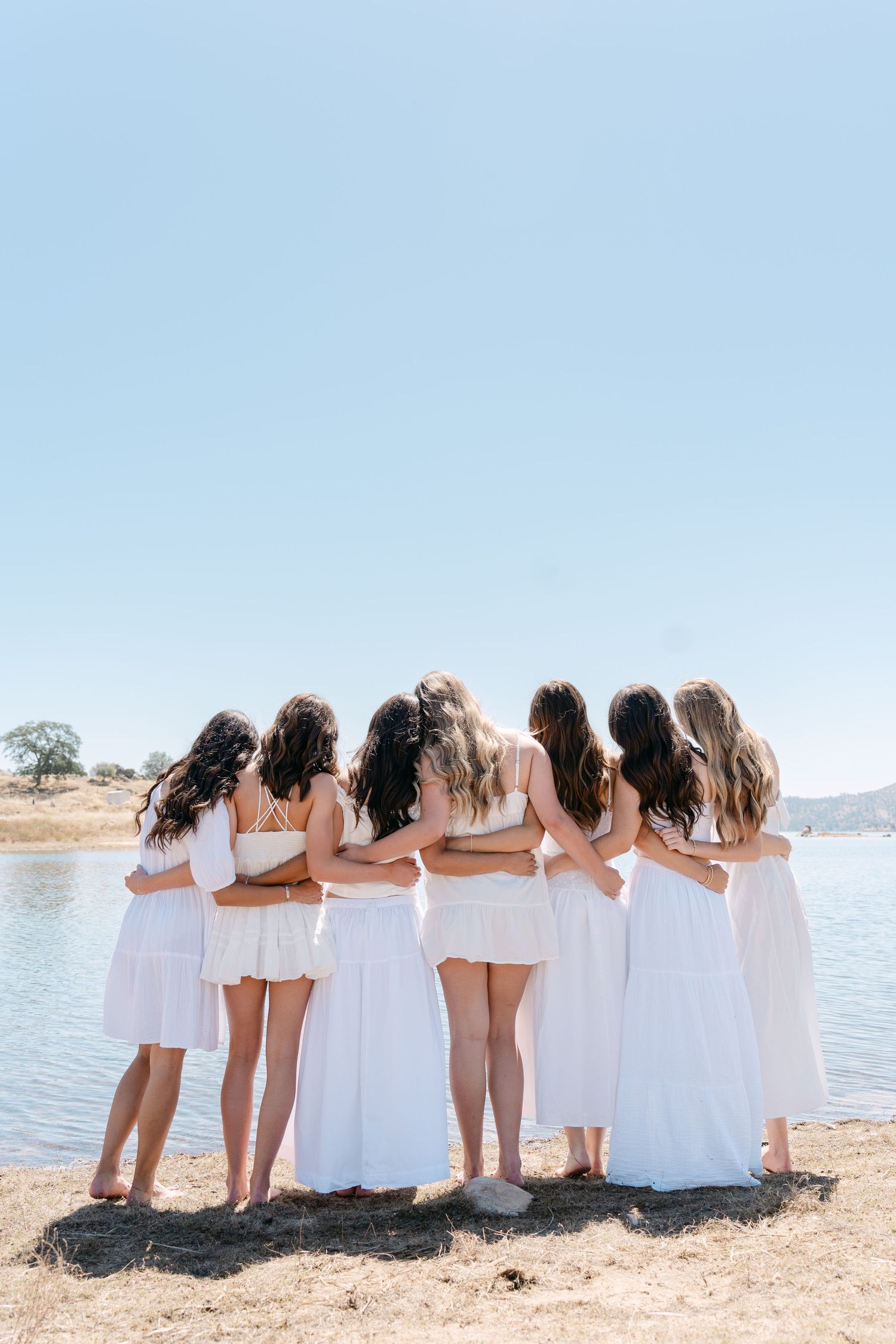 Seven women in white dresses stand arm in arm by a lake, viewed from behind on a sunny day.