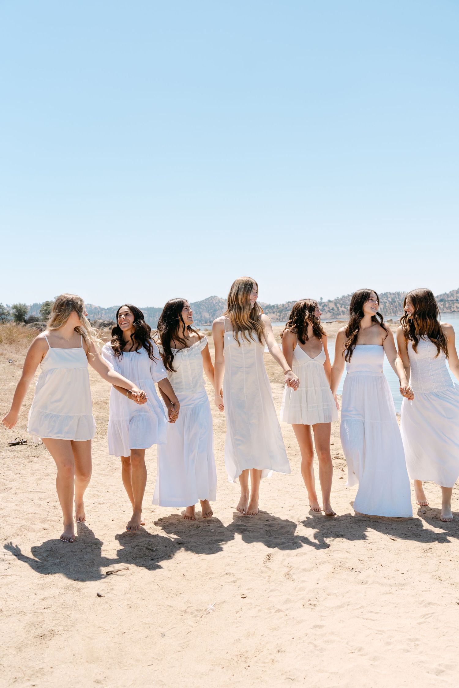 Six women in white dresses walk together on a sandy outdoor setting with hills and clear blue sky in the background.