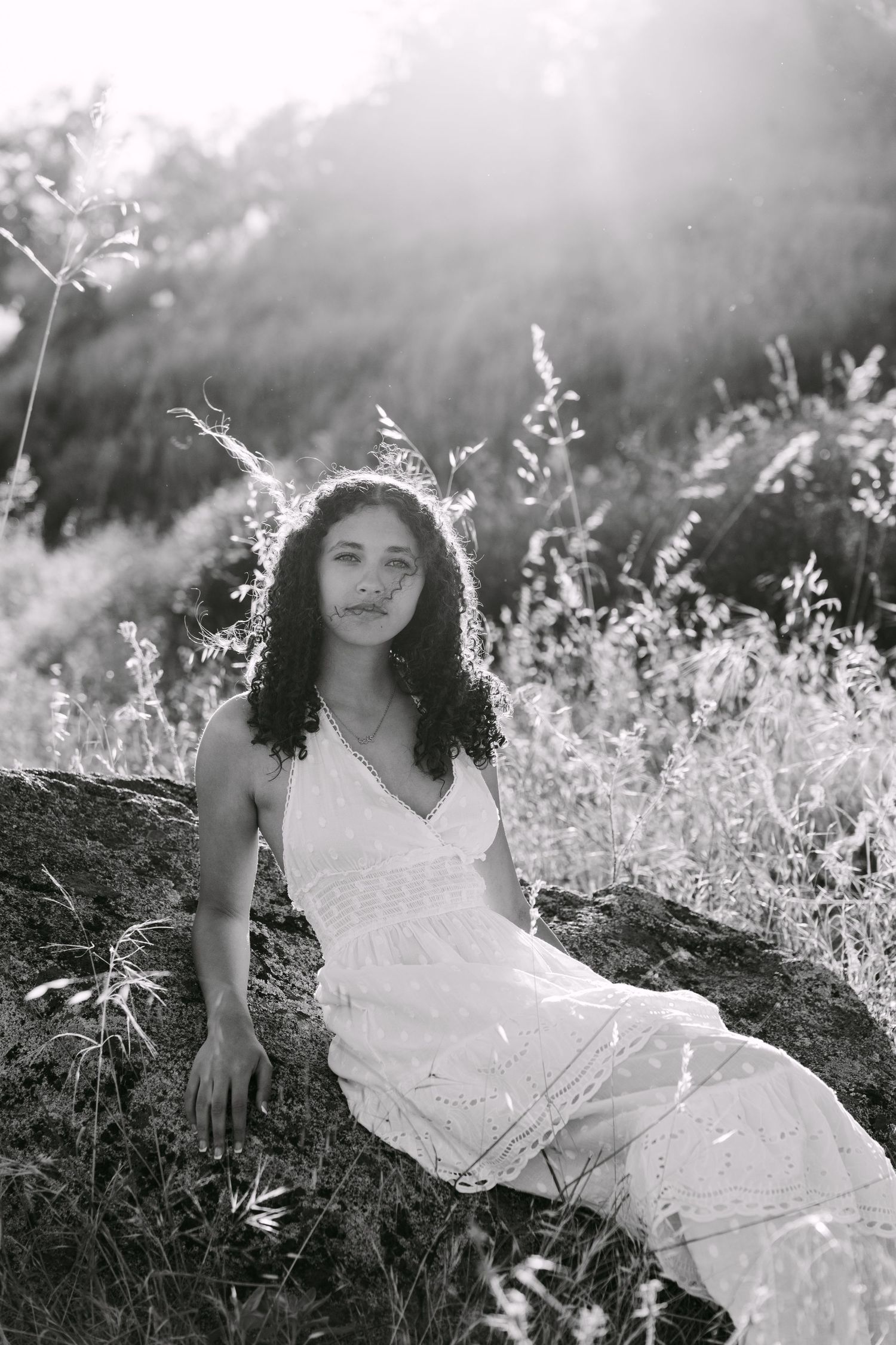 Black and white portrait of a young woman in a white dress sitting on a rock surrounded by wild grass in sunlight.