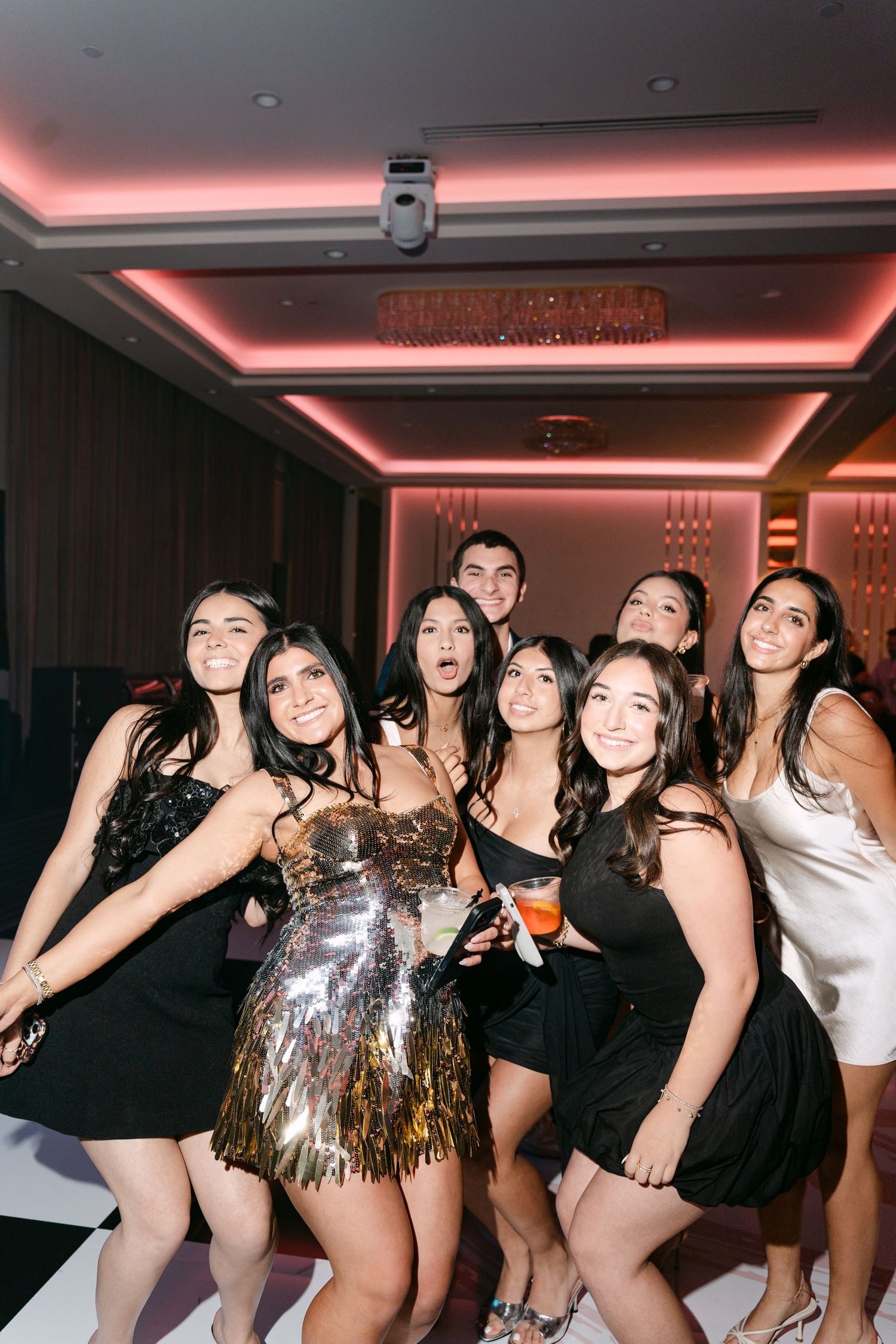 Group of young women in elegant dresses posing together at a formal party event with pink ambient lighting.